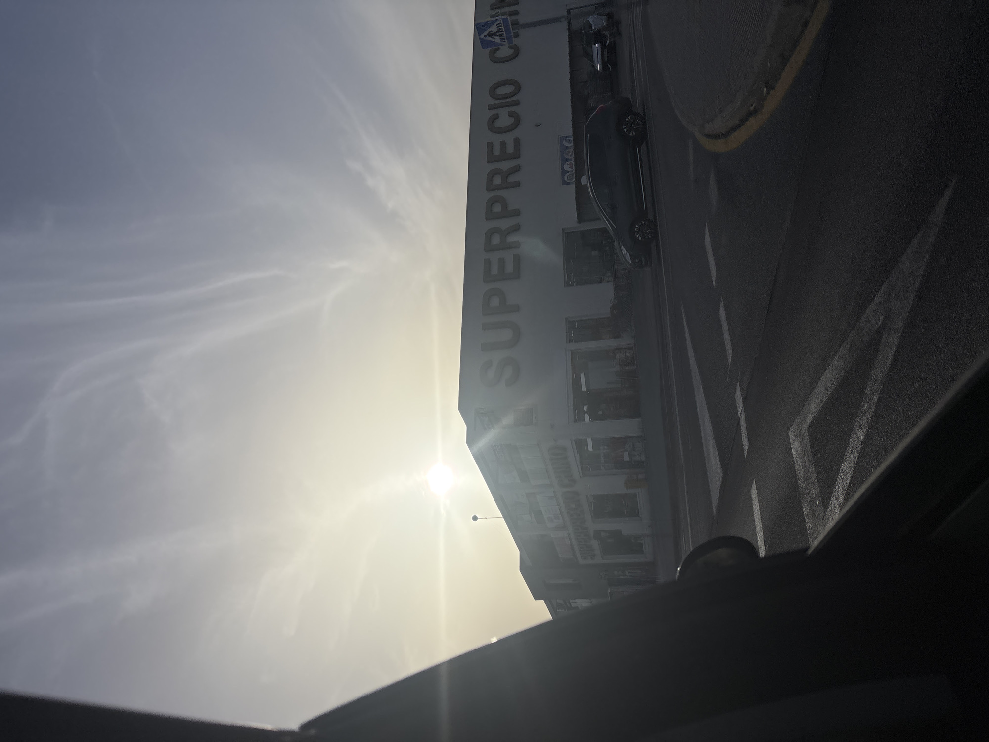 This is a medium-shot, eye-level photograph taken from inside a vehicle, looking out at a street scene in Urbanización La Cenia II, Spain. The sun is low in the sky, creating a bright glare and casting long shadows. A large building with the words "SUPERPRECIO CHINO" emblazoned across its facade dominates the background. Several cars are visible on the road in front of the building, including a dark SUV. The road surface is asphalt with visible white lane markings and a yellow curb. The sky is partly cloudy with wispy formations. The overall lighting suggests it is either early morning or late afternoon.
