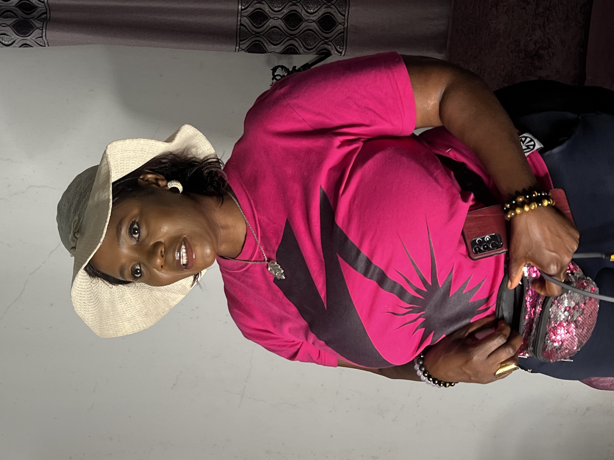 In a medium shot, a Black woman stands indoors, facing left with her head turned towards the viewer. She wears a wide-brimmed, woven straw hat and a bright pink t-shirt with a black graphic print. Around her neck is a silver chain with a leaf-shaped pendant. She has a gold ring on her right index finger and a gold earring in her left ear, shaped like a crescent moon. On her left wrist, she wears several beaded bracelets. She is holding a sequined fanny pack open with both hands, revealing its contents. A smartphone with a dark red case is tucked into her waistband on her right side. The background consists of a plain, textured grey wall and a section of a dark, patterned curtain on the right. The lighting is bright, casting some shadows. The scene appears to be an indoor residential setting. Based on visual cues in the image, the location is a residence in Jos, Nigeria.