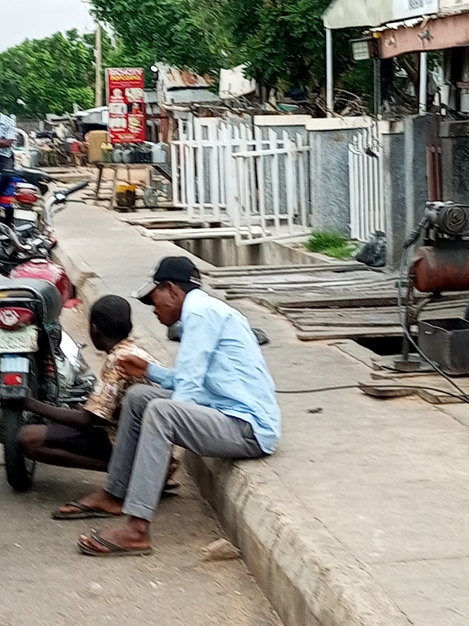 The image depicts an outdoor scene with two individuals on a street in Tempure, Nigeria. In the foreground, two people are seated near a motorcycle. One person, wearing a light blue long-sleeved shirt and grey pants, has a black cap on and is gesturing towards the other person. The second person, a child or youth, is seated next to them, wearing a patterned shirt. They are both wearing flip-flops. The background shows a street environment with a variety of structures, including buildings, stalls, and a white metal fence. There are also some discarded items and equipment visible, such as what appears to be an air compressor. The overall impression is of a busy commercial or street-side area.