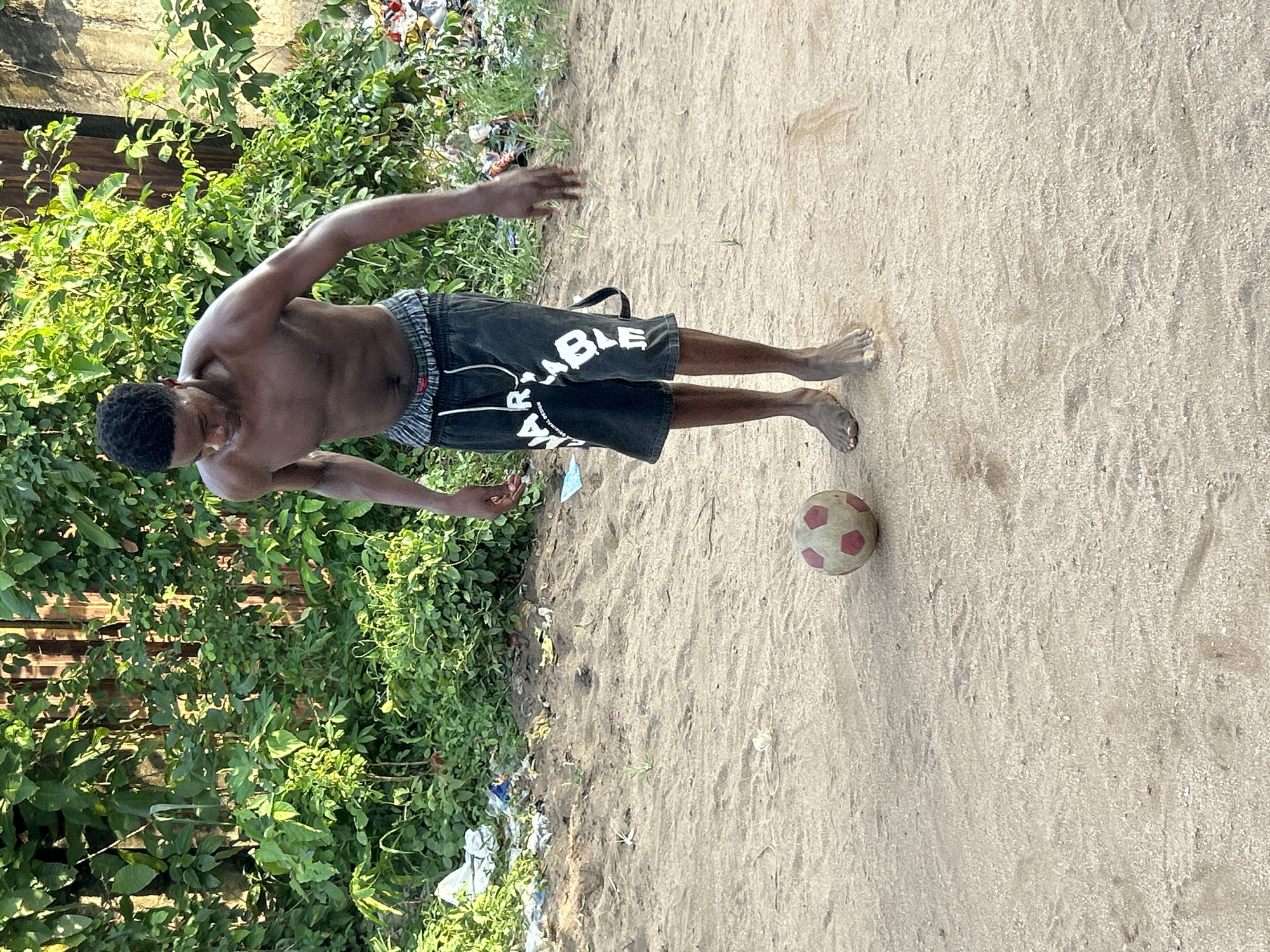 A dark-skinned, shirtless man in dark shorts stands barefoot in the sand, next to a soccer ball, near a lush green area littered with trash. The location is outdoors in what appears to be a beach or sandy area, likely in Ihiagwa, Nigeria based on location data. The man's posture and the presence of the soccer ball suggest he may be playing or about to play. The surrounding environment includes dense vegetation and scattered debris.