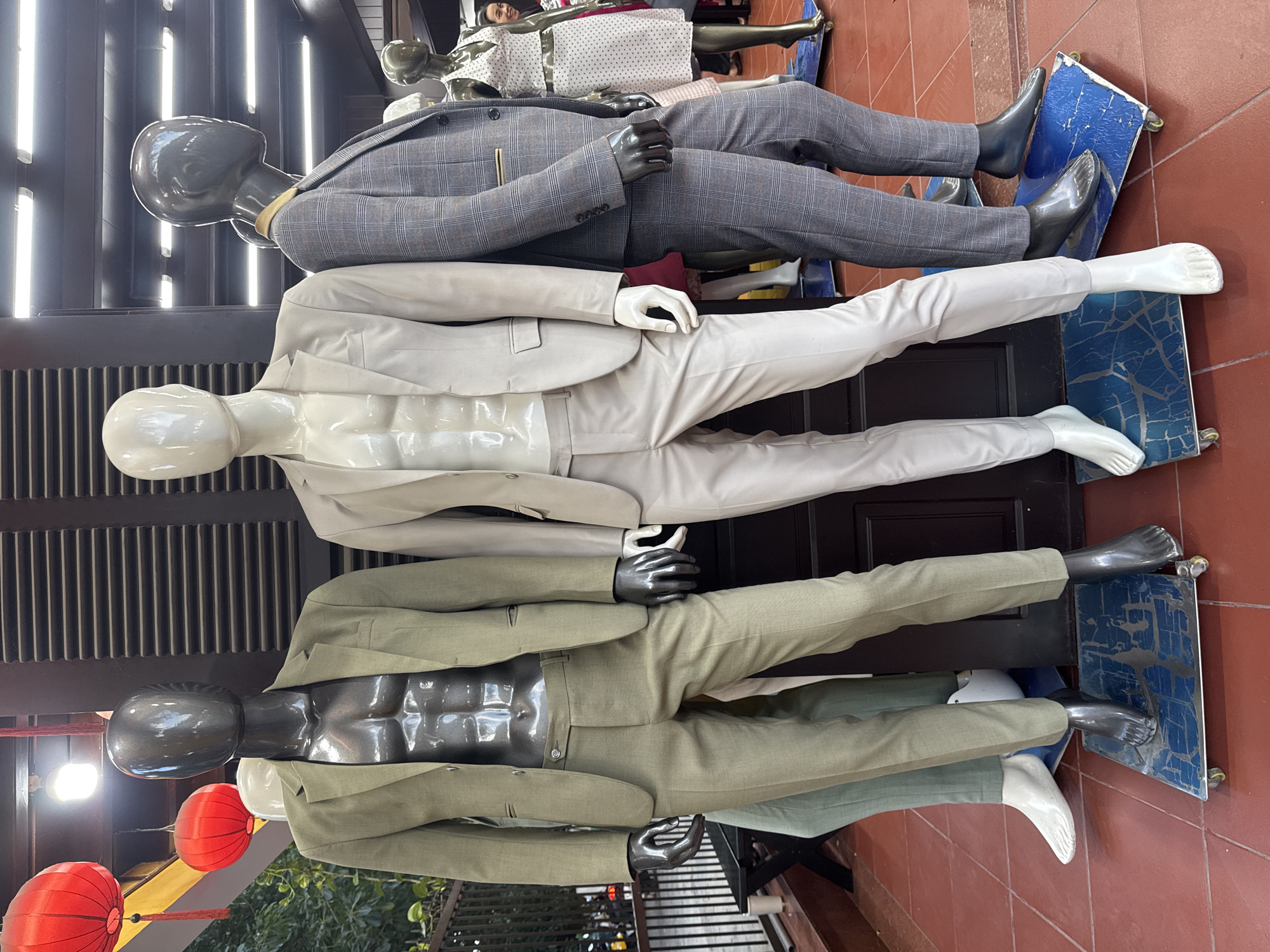 In Hội An Ward, Vietnam, the image shows three mannequins displaying suits. The topmost mannequin wears a grey checkered suit with a yellow lining. Below, a mannequin is dressed in a light beige suit. The bottom mannequin is wearing a green suit. All mannequins are placed on wheeled platforms. Red lanterns hang outside the building visible through a barred window.