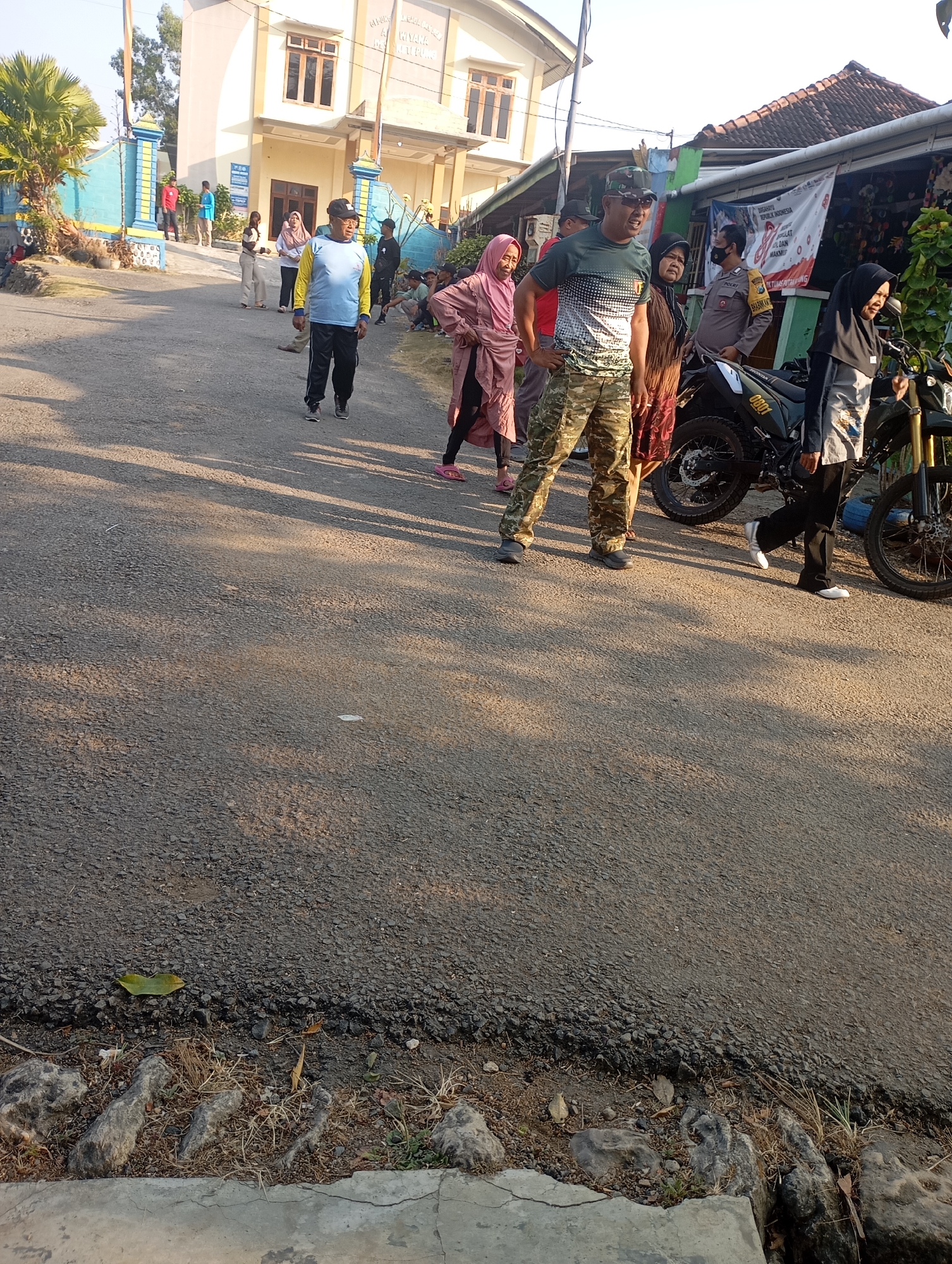 In Ketepung, Indonesia, a group of approximately ten individuals are gathered on an asphalt road in front of a two-story building. The building is light beige with dark brown window frames and a tiled roof. A blue and yellow striped gate is visible to the left of the building. In the foreground, a man in camouflage pants and a green t-shirt with a patterned design is facing left, with his hands on his hips. Beside him, a woman in a brown dress and headscarf stands. To the right of the man in camouflage, a police officer in uniform is standing near a motorcycle. Another woman in a black headscarf and a grey and white patterned top is also near the motorcycle. Several other individuals, including women in headscarves and a man in a blue shirt and black pants, are scattered around the scene, some walking and some standing. The sun casts long shadows across the road, indicating it is likely morning or late afternoon. The environment appears to be a residential or semi-urban area with vegetation visible in the background.