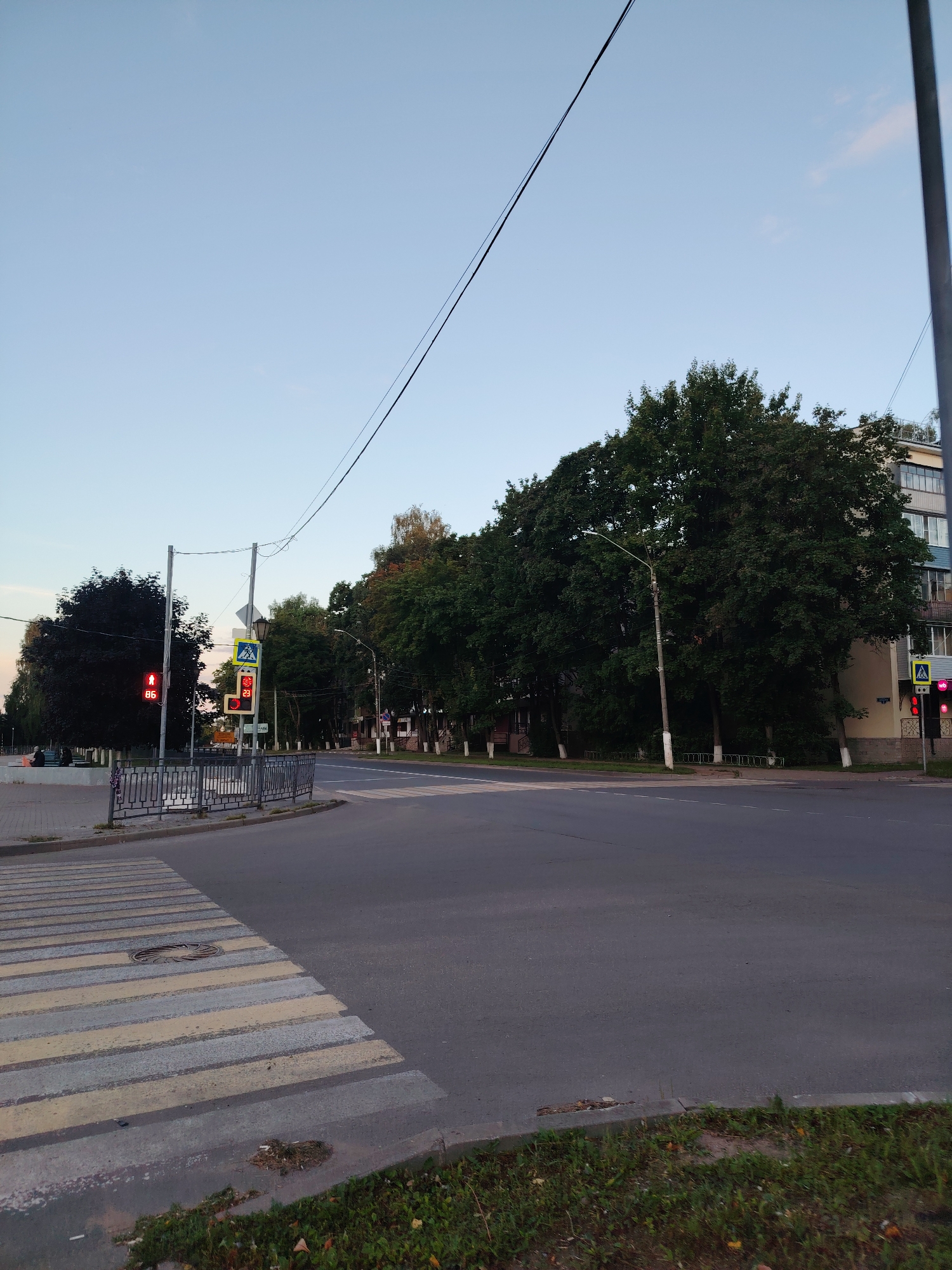 The image shows a street intersection in Konakovo, Russia. On the left, there is a crosswalk with yellow and grey stripes, leading to a sidewalk. A traffic light with a red pedestrian signal and the number "86" is visible. Across the street, there are several large, mature trees lining the sidewalk, with a multi-story building visible behind them. Another traffic signal with a pedestrian crossing symbol is also present. The sky is a clear, pale blue, suggesting it is either early morning or late evening. Power lines are strung across the frame. The overall scene depicts a quiet urban environment.