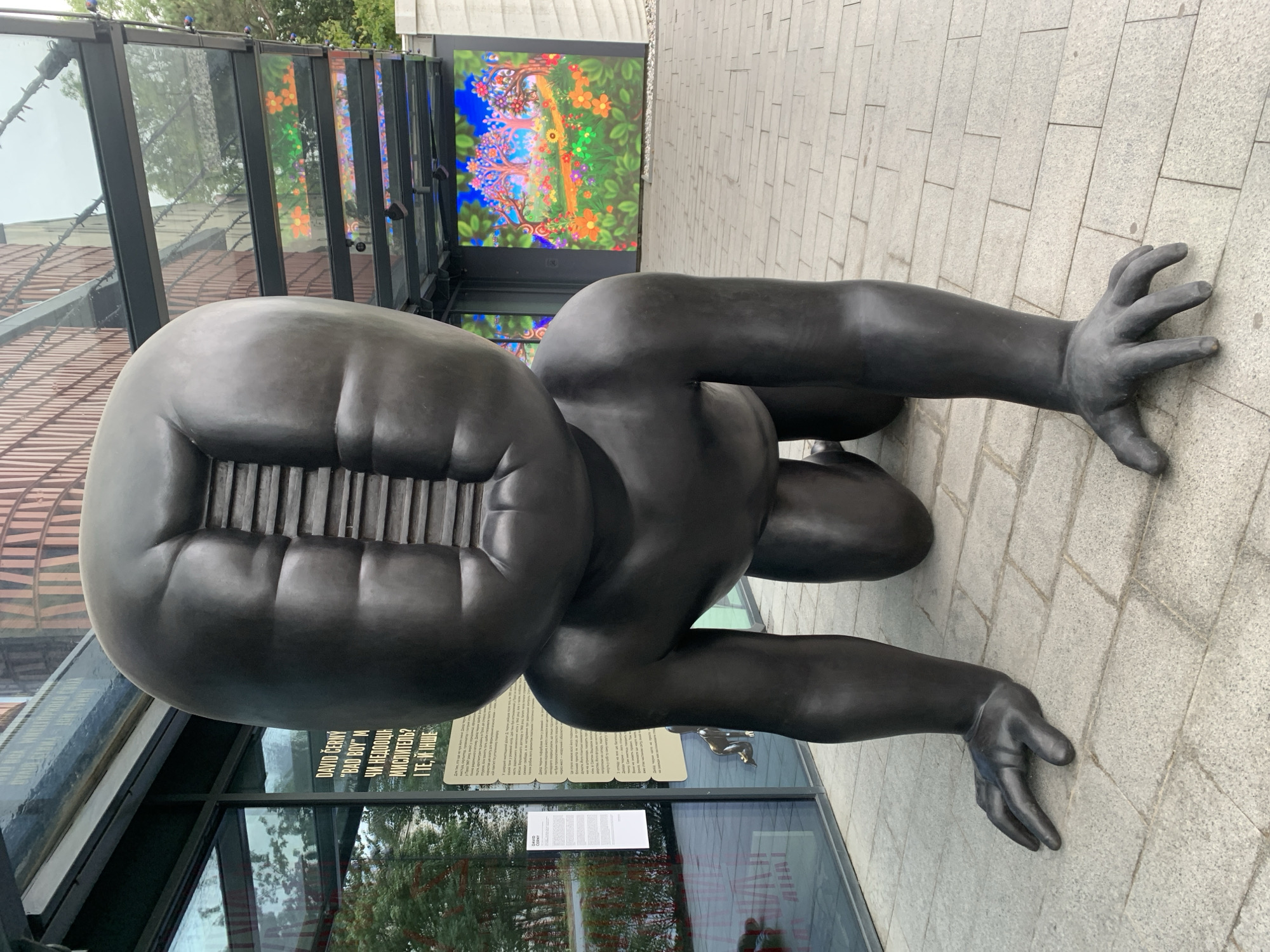 This is a full shot of a large, dark bronze sculpture of a crawling baby with a grate-like opening on its head. The sculpture is on a paved walkway in front of a modern building with large windows. To the left of the sculpture, there is a glass wall with text and images visible behind it. To the right of the sculpture, there is a large, colorful mural of a whimsical landscape with trees and flowers displayed on a wall. The ground is paved with light gray square tiles. The sky is overcast and green trees are visible in the background through the windows. This sculpture, "Rider" by David Černý, is located in front of the "Fabrika" cultural center in Lviv, Ukraine.