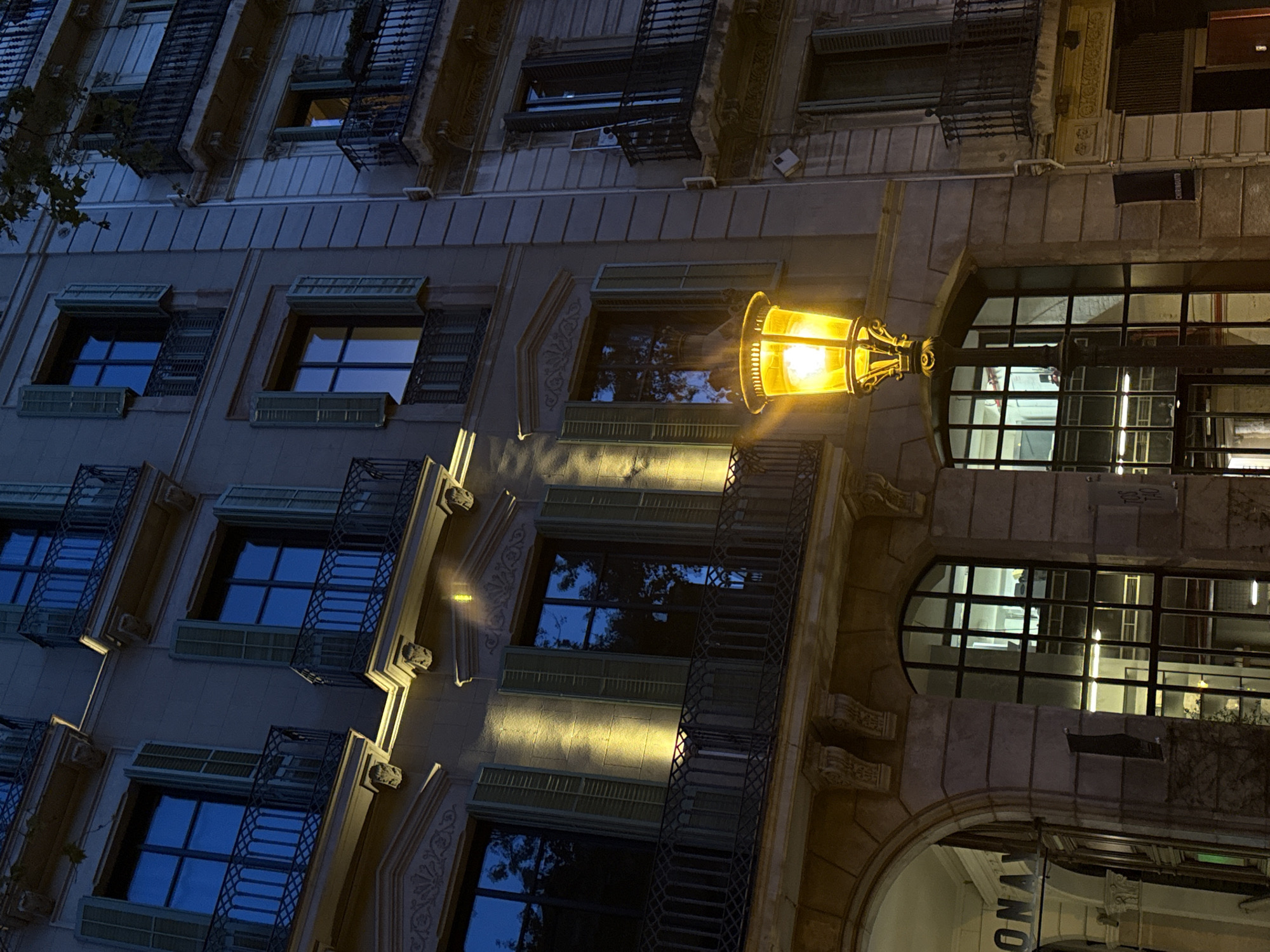 The image captures the facade of a building in Barcelona, Spain at night, illuminated by a street lamp. The building is characterized by a row of windows, some with shutters and ornate iron balconies. The windows reflect the dim blue light of the night sky. A prominent street lamp with a traditional design casts a bright yellow glow onto the building, creating a contrast between the warm light it emits and the cool tones of the night. At the bottom right, a partial view of an arched entrance is visible, with signage, suggesting a commercial establishment. The overall setting conveys an urban, nighttime scene in a historic part of Barcelona.