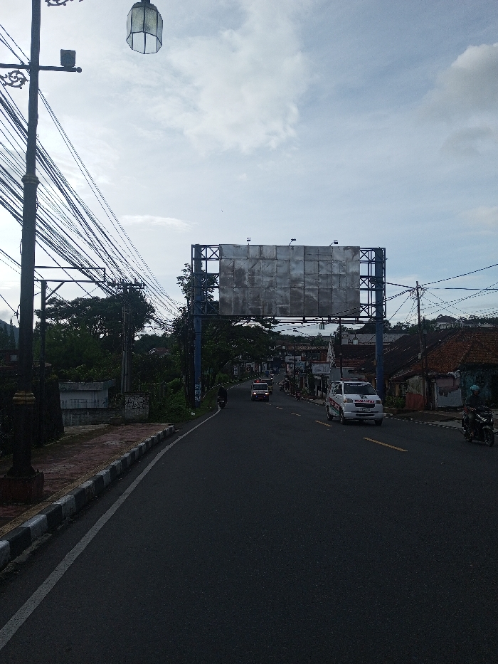 A road stretches forward beneath an overcast sky. Utility poles laden with wires line the left side of the road. A large, blank billboard spans the road ahead. Vehicles, including an ambulance, are visible driving in the distance. A curb runs along the left edge of the road. A streetlamp stands tall in the left foreground.