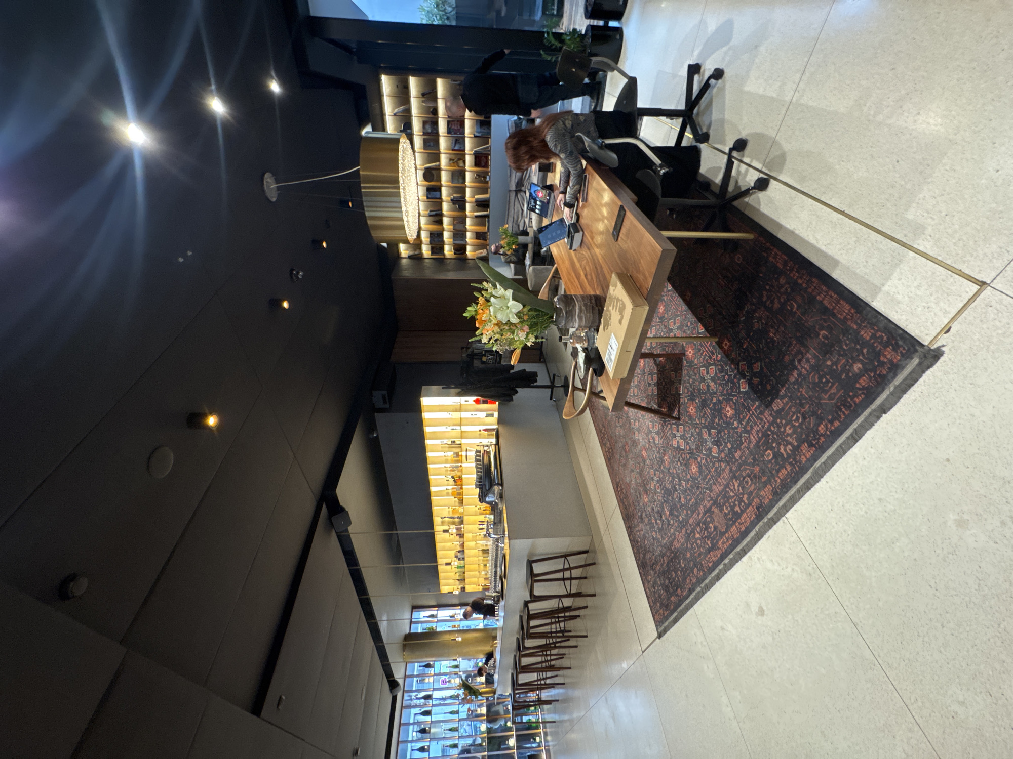 The image depicts the interior of the Sense Hotel Sofia Lobby Bar & Library in Sofia, Bulgaria. The scene is illuminated by artificial lighting, including warm yellow backlighting behind two bar areas and overhead recessed lights, with a dark-colored ceiling. The flooring consists of light-colored terrazzo or polished concrete, partially covered by a large, dark red and blue ornate rug.
On the left, a long, light-toned bar counter is fronted by seven wooden bar stools with dark seats. Behind the counter, an illuminated display showcases numerous liquor bottles on shelves, complemented by an adjacent glass wall exhibiting additional bottles. Two individuals are present behind the bar; one is standing further down, and another is partially visible near an espresso machine and taps.
In the center-right, a large, rectangular wooden table is occupied by a person with reddish-brown hair, seated in a black office chair, actively using a silver laptop and a secondary screen. A vase containing a prominent floral arrangement of white, orange, and green flowers, a small cardboard box, and a dark bottle are on this table. A smaller wooden side table with brass legs is positioned nearby on the rug. Overhead, a large, circular gold-colored chandelier hangs.
Behind the main table, a dark wall features shelves displaying numerous books or objects, lit from behind. Further back, a man in dark attire is standing near a large window or glass door, facing away from the viewer. A grey sofa and a coffee table are visible beyond the main table, with another person partially obscured, possibly engaged with a device. A black coat rack with clothing is present on the far right, near a potted plant.