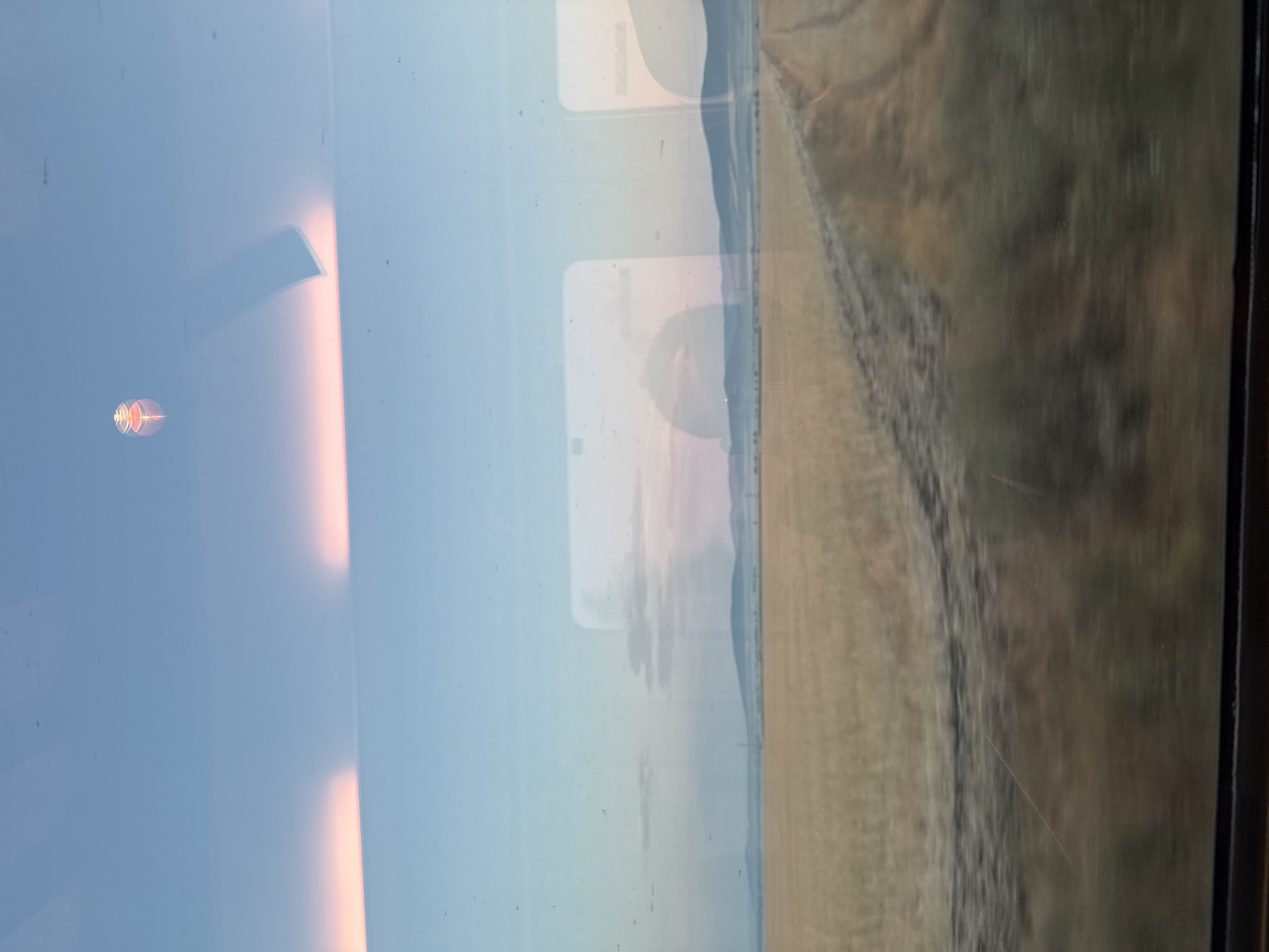The image shows a view from inside a vehicle, looking out a window at a landscape. The sky is a pale blue with soft pinkish-orange clouds near the horizon. A faint reflection of what appears to be interior lights and a small, abstract red and yellow object are visible on the window. Below the sky, a vast field of dry, golden grass or crops stretches towards distant, hazy blue hills. A dirt road or track cuts through the field diagonally from the right side of the frame, leading towards the hills. There are also indistinct reflections of the vehicle's interior on the window, including rectangular shapes that suggest other windows or panels. The overall impression is of a rural landscape viewed during twilight or dawn. There are no people or discernible activity in the scene. The provided information about Chernograd, Bulgaria, does not directly correlate with any visual elements in the image itself.