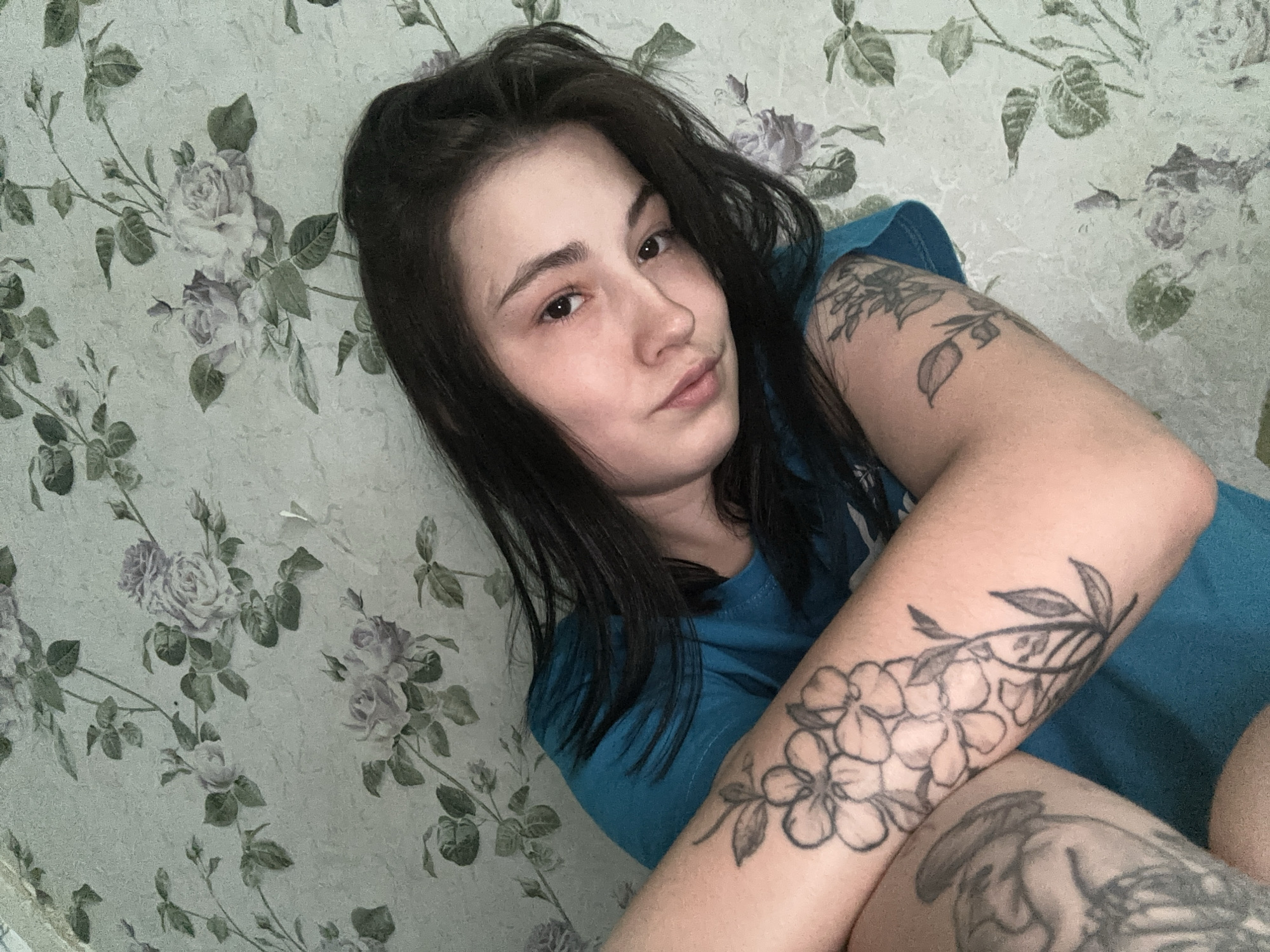 A young woman is lying down against a floral patterned wall. She has dark hair and light skin. She is wearing a blue shirt and has several tattoos on her arm. The floral pattern on the wall is primarily shades of grey and green. The setting is unknown.