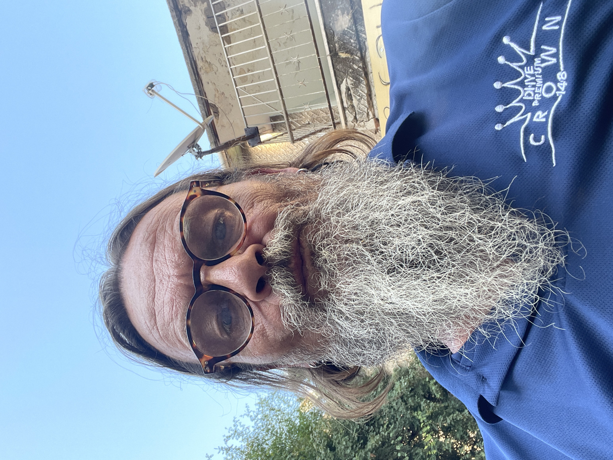 This is a vertical, close-up, selfie-style image of a person with a long, gray beard and long, brown hair. The person is wearing glasses with tortoiseshell frames. They are also wearing a blue collared shirt with an embroidered white logo on the chest that reads "CROWN" with "DHVE PREMIUM" above it and "148" to the right.
The background shows a clear blue sky, a portion of a building with peeling paint and a metal grate, and some green trees. A satellite dish is visible on the wall of the building. The lighting suggests it is daytime. The overall impression is of a casual outdoor setting, possibly in a residential area, given the building and satellite dish. The image is shot from a low angle, looking up towards the sky.