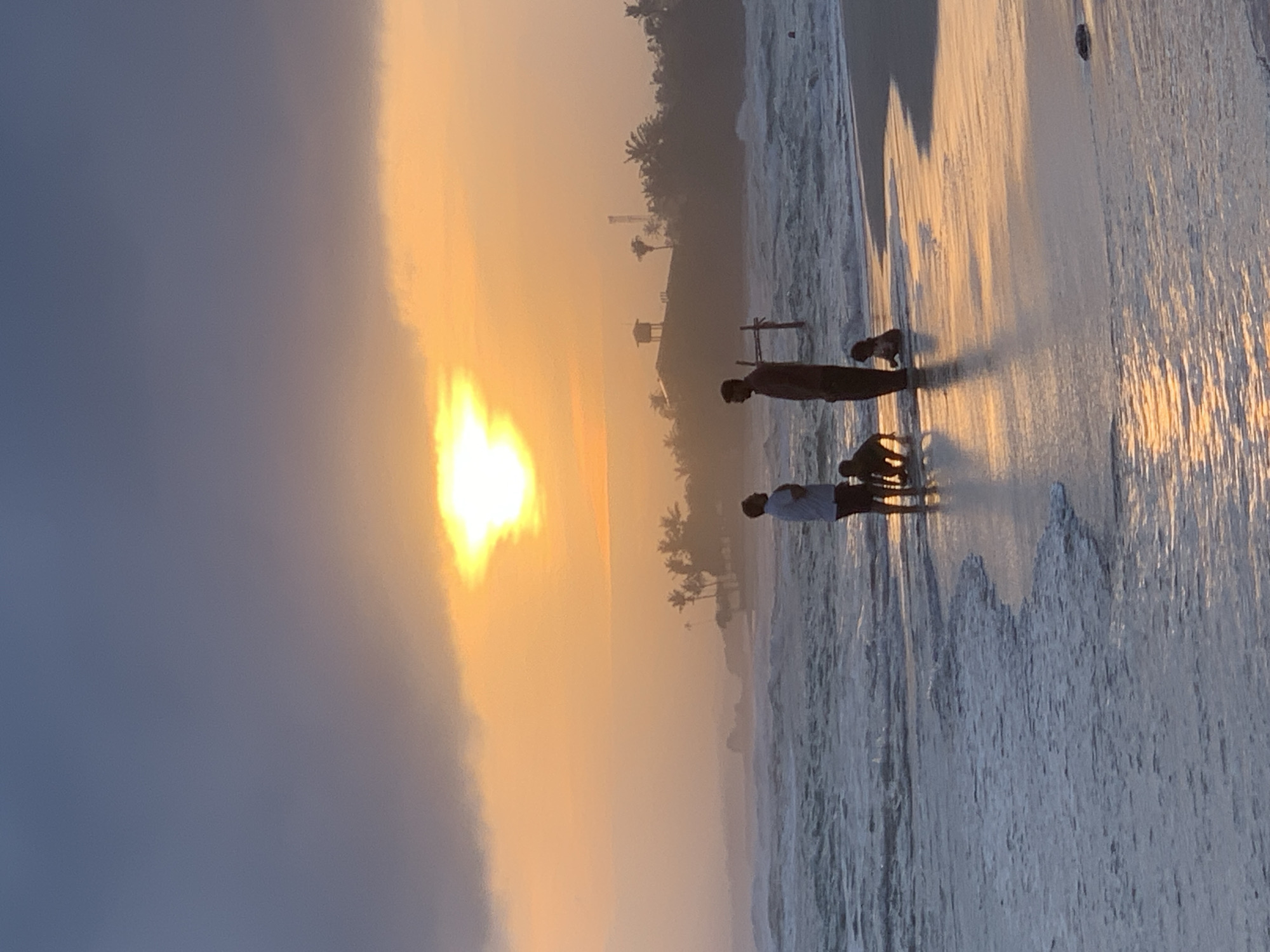 This image depicts a serene sunset scene on the beach in Kathaluwa, Sri Lanka. In the foreground, the gentle waves of the ocean lap the shore, reflecting the warm hues of the setting sun. Three individuals and two dogs are silhouetted against the vibrant sky. Two adults, one standing and the other bent over, are interacting with two young children who are playing near the water's edge. The backdrop features a hazy coastline dotted with palm trees, and a building with a water tower. The sky is a canvas of dramatic colors, with dark clouds parting to reveal a brilliant sun, casting a golden glow over the entire scene. The overall atmosphere is one of tranquility and family enjoyment by the sea.