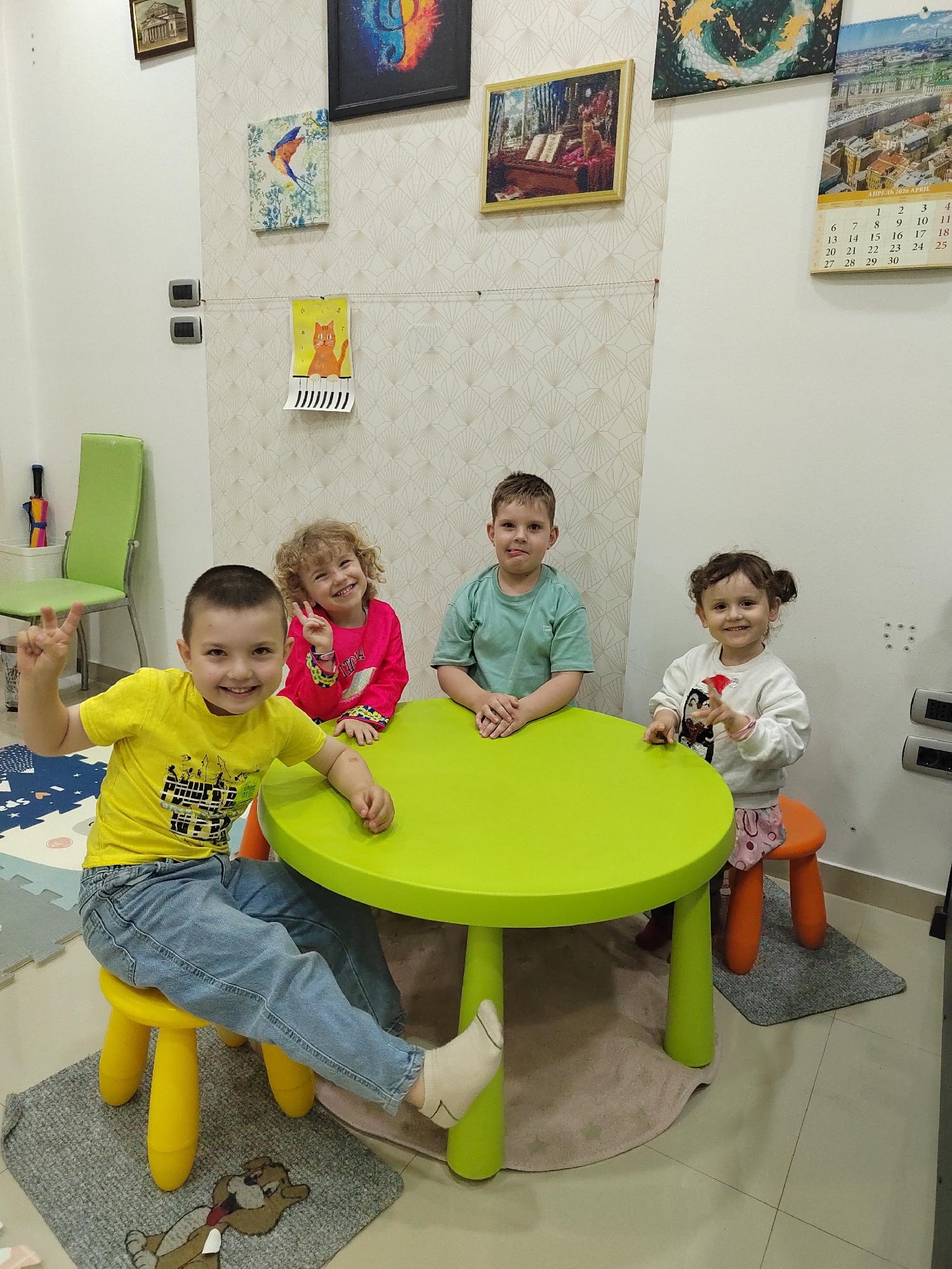 Four young children are seated around a bright lime-green, round table in what appears to be a brightly lit indoor playroom in Podgorica, Montenegro. All children are smiling broadly at the camera, conveying a happy and playful atmosphere.
To the left, a boy in a yellow t-shirt and blue jeans makes a peace sign with his right hand while seated on a yellow stool. Beside him, a girl with curly blonde hair, wearing a pink long-sleeved top, also flashes a peace sign. Opposite them, another boy in a light green t-shirt rests his arms on the table. To his right, a girl with dark hair tied in pigtails, dressed in a white long-sleeved top and patterned bottoms, points with her finger while sitting on an orange stool.
The background features white walls, partly covered with patterned beige wallpaper. Various artworks adorn the walls, including framed pictures of a classical building, an abstract piece, a bird on a branch, and a cat playing an organ. A child's drawing of an orange cat playing a piano is strung across the wall with small clothespins. A calendar is visible on the right, displaying "АПРЕЛЬ 2026 APRIL" and a grid of dates from 1 to 30, with "MON" partially visible. A green chair, a multi-colored umbrella, and several electrical outlets are also present in the room. Playmats and small rugs, including one with a cartoon character, are visible on the floor beneath the table.