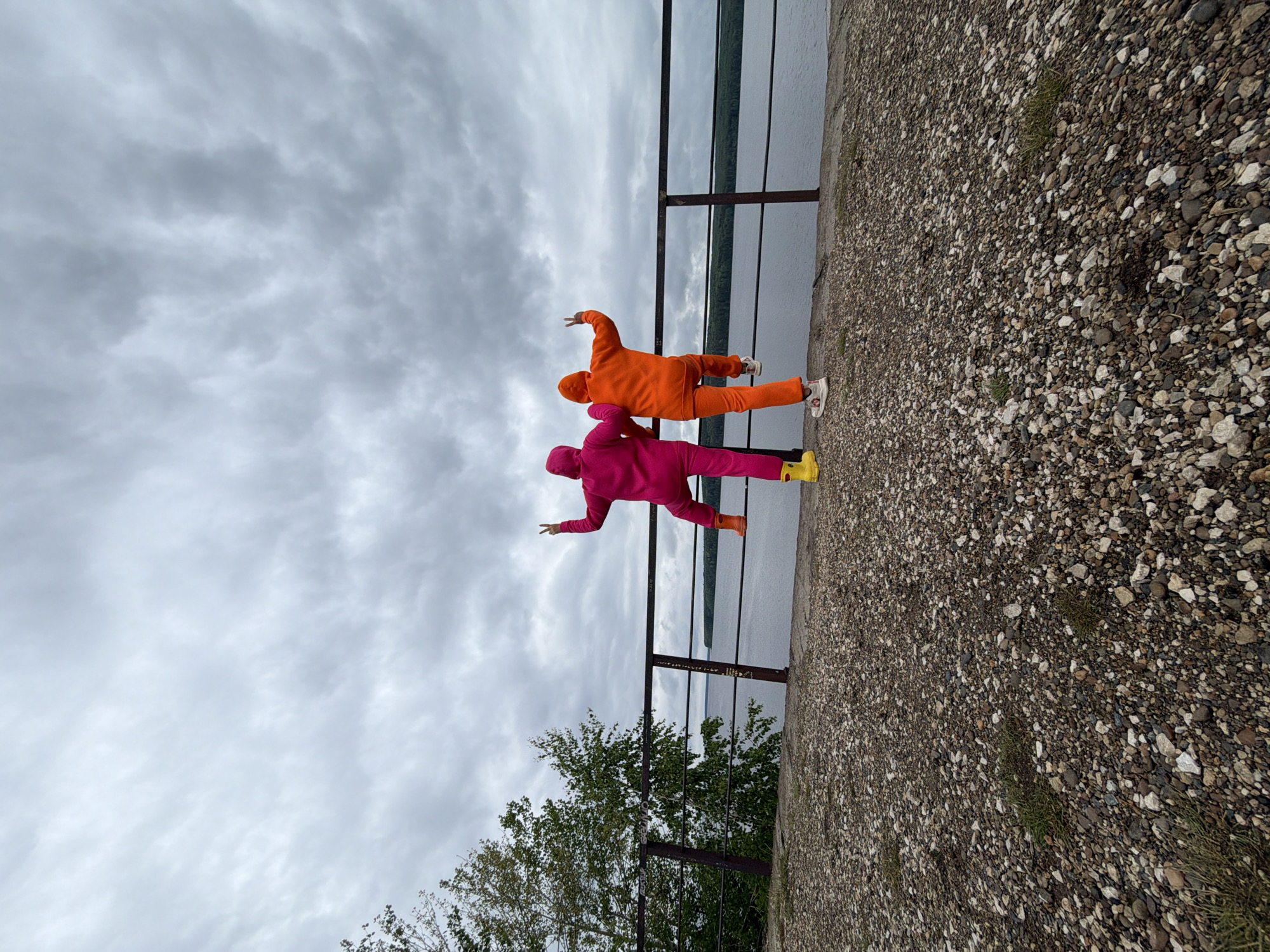 Two individuals, one in an orange sweatsuit and the other in a pink sweatsuit, stand on a paved embankment in Chaikovsky, Russia, with a metal railing separating them from a body of water. The embankment's surface transitions into a gravel area. The sky is overcast. The individuals strike playful poses.