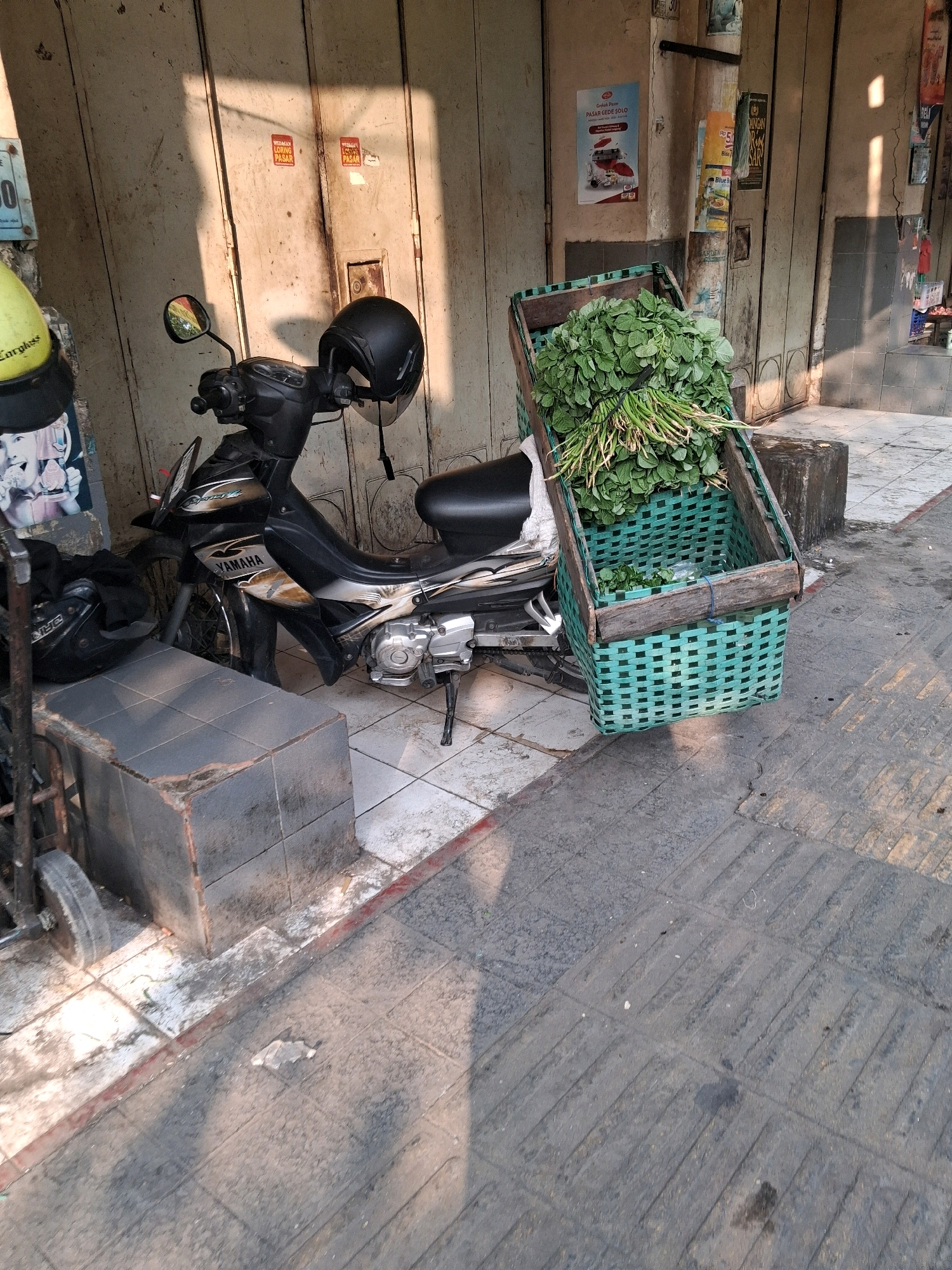 A motorcycle is parked next to a stall in Surakarta, Indonesia. The motorcycle is black and gold with "YAMAHA" written on its side. A black helmet rests on the handlebars. In front of the motorcycle is a large green woven basket filled with leafy green vegetables. The basket is wooden on top and attached to the motorcycle. The stall has a weathered wooden door and some posters are taped to it. The ground is paved with grey tiles and concrete. Sunlight casts shadows on the scene.