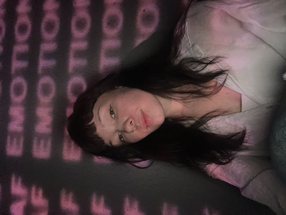 A young woman with dark hair and bangs lies in a dimly lit space. The word "EMOTION" is projected in pink onto the wall behind her, creating a repetitive pattern. She wears a light-colored, button-down shirt and has a nose ring. She looks towards the camera with a slight smile. Her skin is fair, and her hair is softly styled around her face. The overall mood is intimate and relaxed, with a touch of artistic flair.