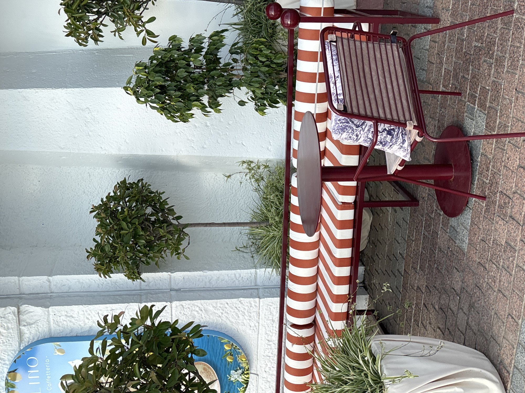 An outdoor seating area features a long, striped bench with red and white cushions and a small, round, red table. A single red chair with a patterned cushion sits next to the table. Green plants are visible in the background and in a large white planter to the left. The scene is set on a brick-paved area in front of a white, textured wall. A blue sign with text and an image of lemons is mounted on the wall. The overall impression is of a cafe or restaurant setting.