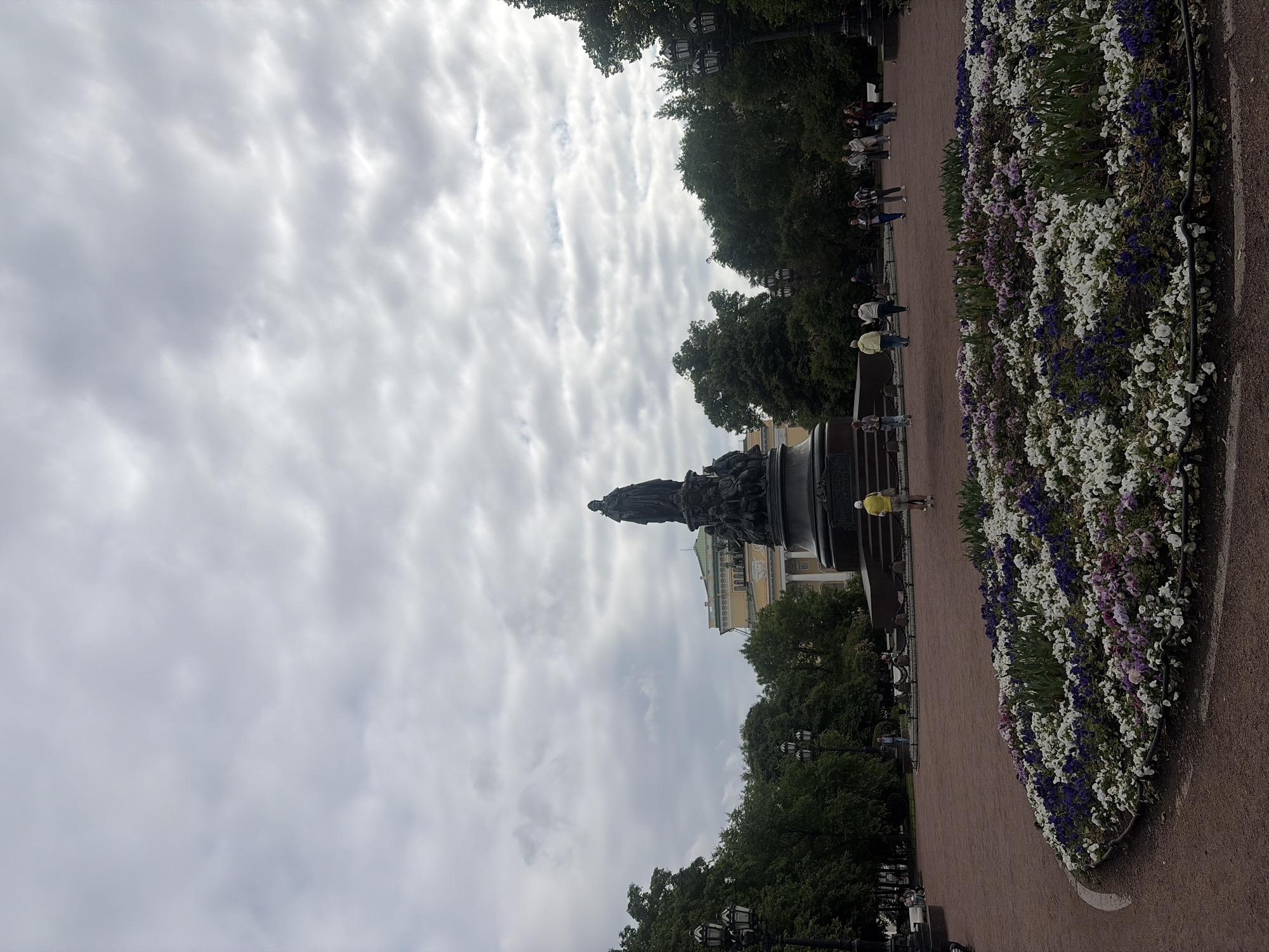 The image depicts a cloudy day at Palace Square in Saint Petersburg, Russia, featuring the Monument to Nicholas I. A large bronze statue of Nicholas I on horseback dominates the center of the composition, positioned on a substantial granite pedestal. In the foreground, a circular flowerbed is filled with a vibrant mix of white, purple, and pink flowers. Several people are visible walking around the square and near the monument. To the left and right of the monument, trees provide a green backdrop, and lampposts are scattered throughout the park-like setting. In the background, the pale yellow facade of a building, likely the General Staff Building, is partially visible. The ground is covered with a reddish-brown gravel path.