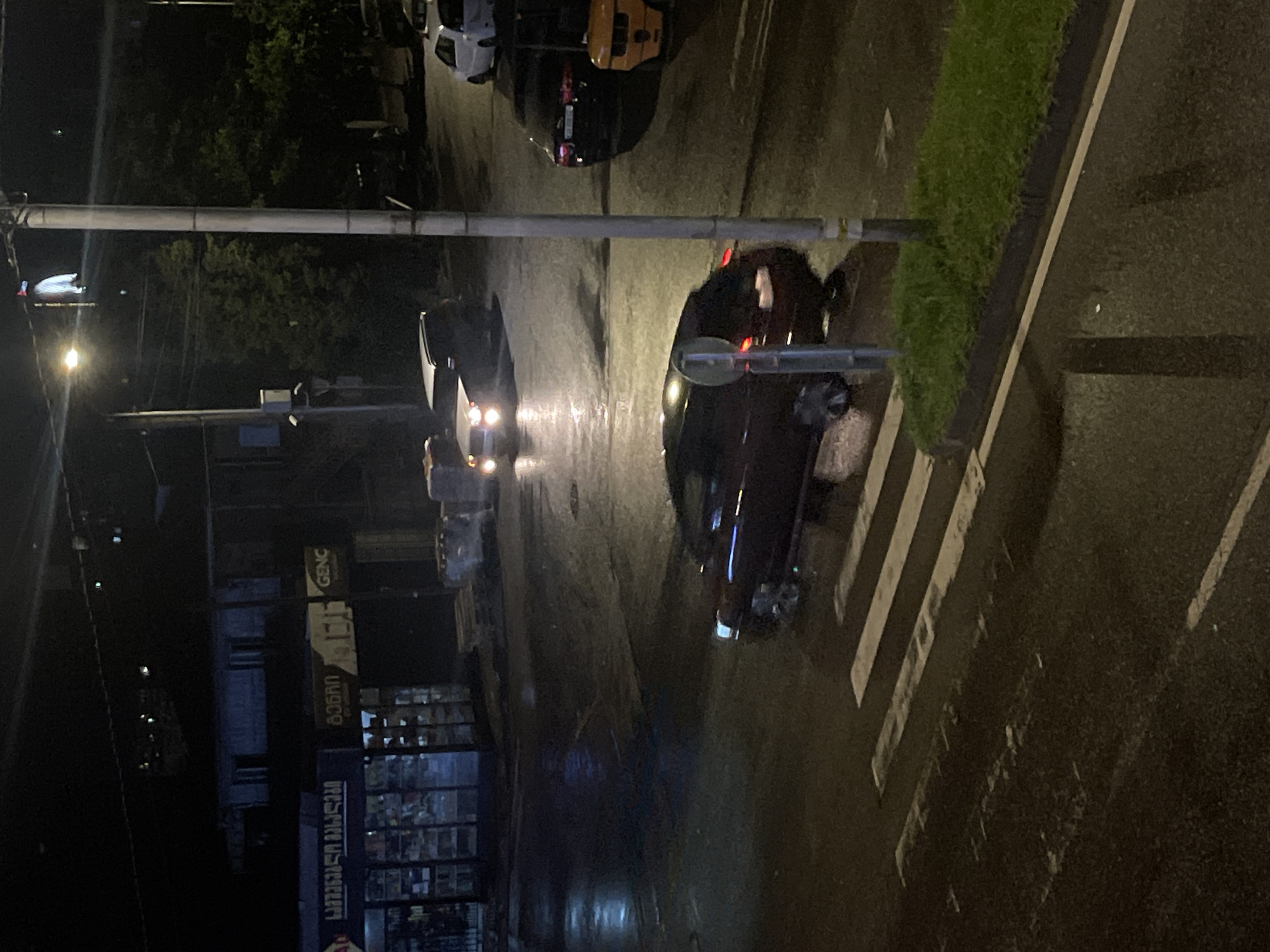 The image captures a nighttime street scene in Batumi, Georgia. The ground is wet, indicating recent rain, and reflections of lights are visible on the asphalt. In the foreground, a crosswalk with white stripes is present. To the right of the crosswalk, there is a strip of green grass with a dark curb.
Several vehicles are present in the scene. A dark-colored car is positioned on the crosswalk, facing towards the right side of the frame. Further back, a white SUV with its headlights on is visible in the intersection, also appearing to be moving. In the background, to the right, a black car is parked or stopped. Another yellow vehicle, possibly a small construction vehicle or utility cart, is partially visible.
A lamppost stands prominently in the mid-right of the image. In the background, a building with multiple windows and a sign that appears to say "GENC" is dimly lit. A street sign is also visible on a pole. There are no visible people in the frame. The overall atmosphere is dark and somewhat moody due to the nighttime lighting and wet conditions.
