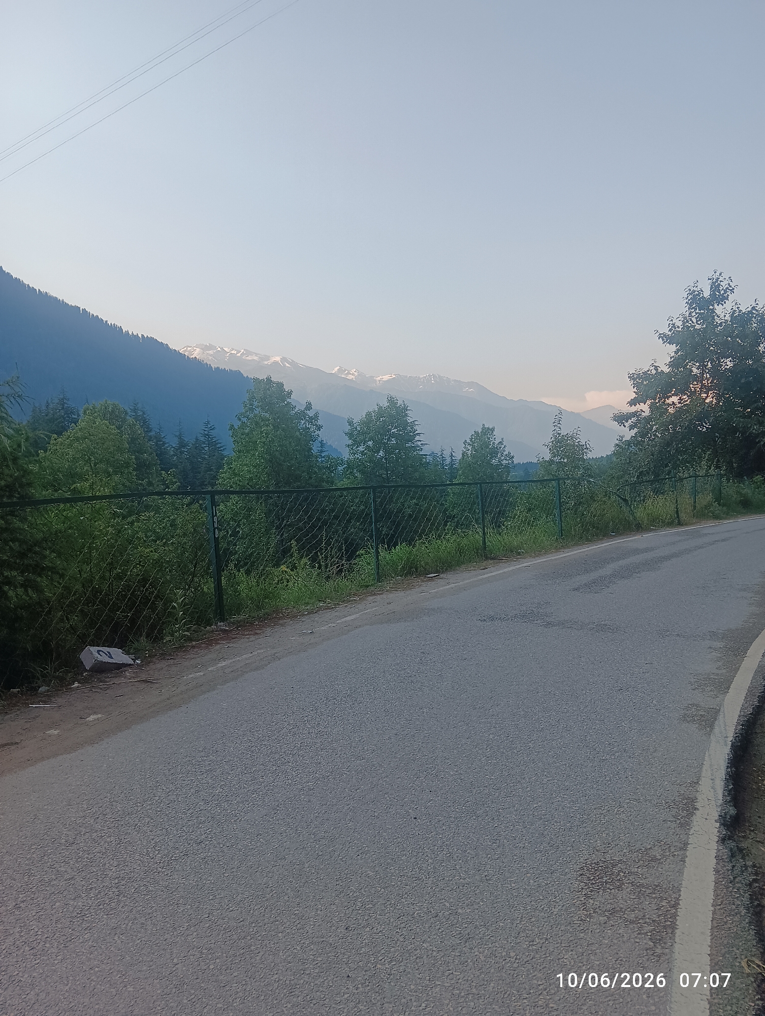 A paved road curves to the right, bordered by a chain-link fence and lush green foliage. Beyond the fence, a dense forest of pine trees covers a steep mountainside, its peak dusted with snow. The sky above is a clear, pale blue, with three thin power lines stretching across it. The scene is lit by the soft glow of early morning light, as indicated by the timestamp "10/06/2026 07:07" in the lower right corner of the image. The image captures a serene natural landscape on the outskirts of Manali, India.