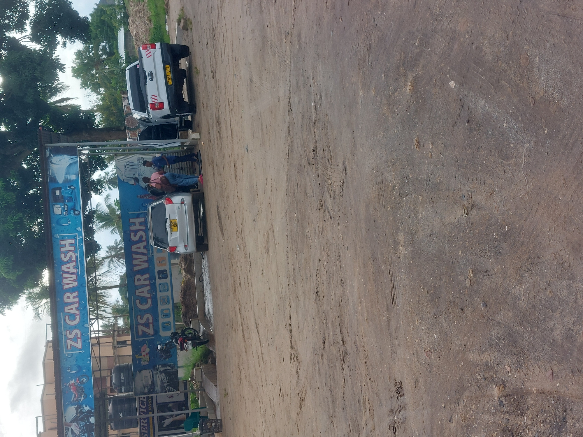 The image depicts an outdoor scene at "ZS Car Wash," likely located in Dar es-Salaam, Tanzania, based on Tanzanian license plates observed on the vehicles. Two vehicles are present, a white pickup truck and a white SUV. Several individuals appear to be interacting near the SUV, with one person in a pink shirt visible. The ground is primarily dirt, with tire tracks and some gravel visible. Vegetation is present on the left side of the frame, with trees and foliage visible. The car wash's signage is prominently displayed with blue and white colors and the text "ZS CAR WASH" written on it. A motorbike is parked near the signage. There are water tanks and cleaning equipment suggesting operation of a car wash at the scene.