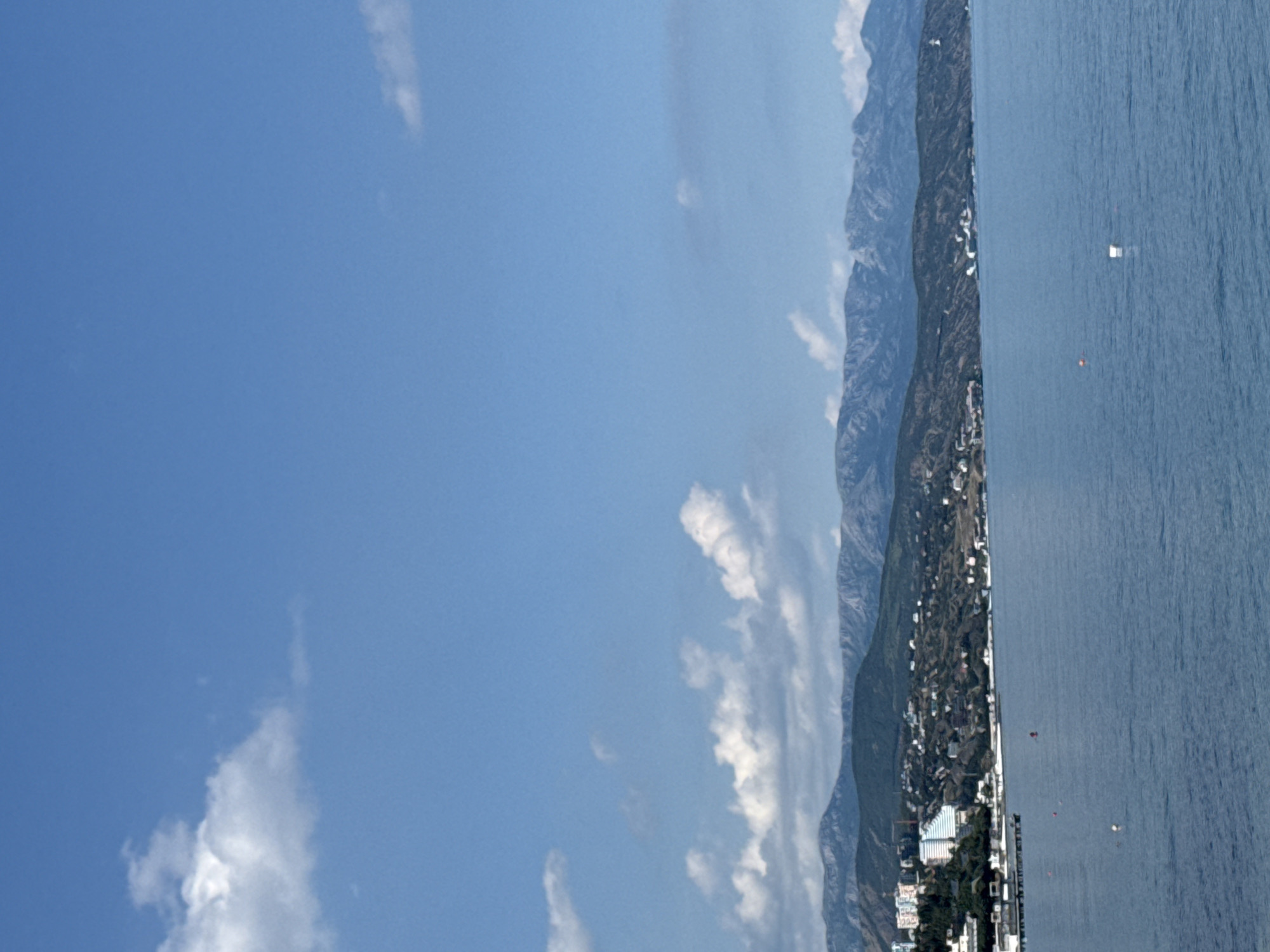 The image displays a vertically oriented panoramic view of a coastal landscape. The left half of the frame is dominated by a clear, bright blue sky with scattered white clouds; some are wispy, while others are more defined cumulus formations.
On the right side, a prominent mountain range runs vertically, forming a rugged backdrop for a coastal settlement. These mountains are covered in dark green vegetation, suggesting a forested environment. A narrow strip of land is situated between the mountains and the sea, featuring a densely built-up area with numerous buildings, indicating a town or city. Various structures, from smaller buildings to larger multi-story developments, are visible, interspersed with green areas and trees.
The far-right portion of the image is occupied by a vast body of deep blue water, presumably the sea or an ocean. The water's surface shows fine textures, suggesting calm ripples or small waves. Several small objects are present in the water: a distinct white rectangular object (possibly a boat or a floating platform) in the mid-right, and closer to the shore, a few indistinct colorful buoys or markers (one orange, one yellow). Another small white object is visible further out.
The scene depicts a sunny day with clear skies and excellent visibility, indicative of late morning or early afternoon lighting. The overall impression is one of a serene coastal environment with a settled community. No specific human activities are clearly depicted, nor is any text visible in the image.