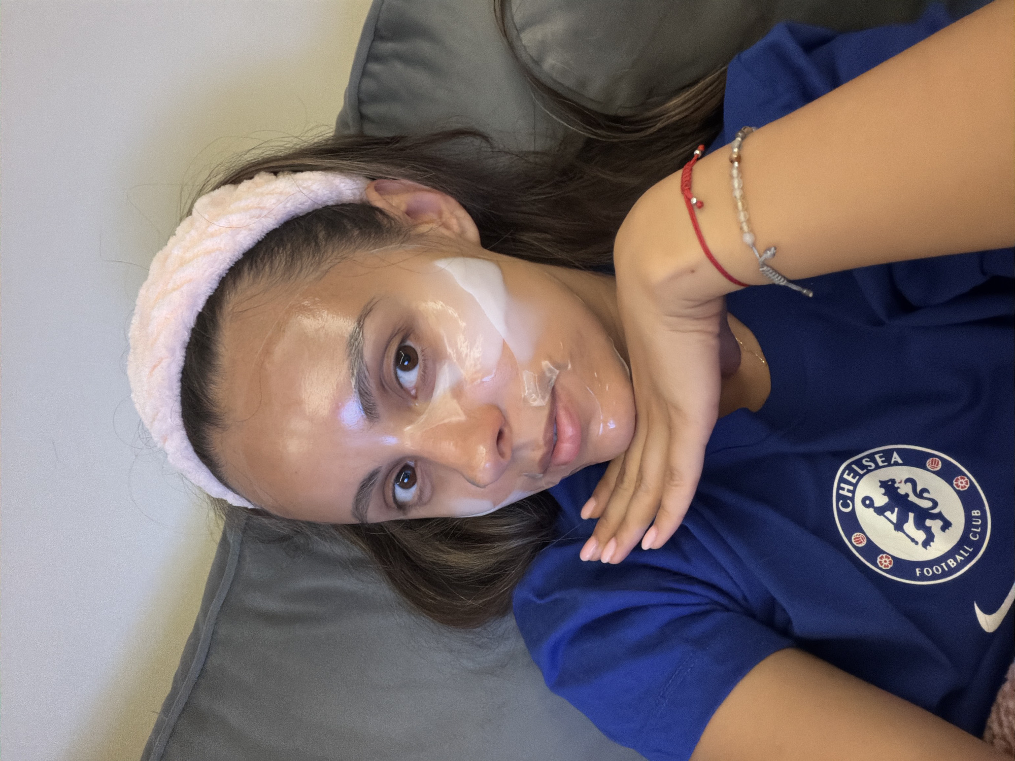 A young woman with a facial mask is lying down on a gray, possibly velvet, couch. She is wearing a pink headband and a blue t-shirt with the Chelsea Football Club logo. Her right hand is raised towards her face, adorned with a red thread bracelet and another beaded bracelet. The background is a plain, light-colored wall. The location is unknown.