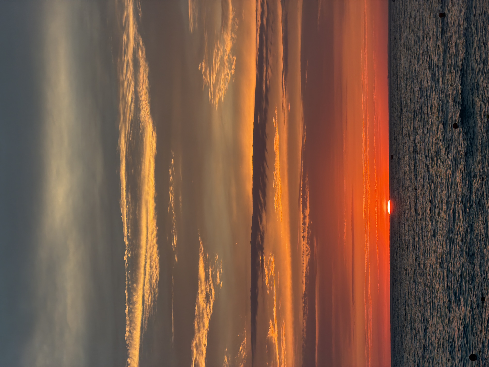 The image depicts a sunset over the Black Sea, viewed from the city of Sochi, Russia. The sky is filled with vibrant hues of orange, red, and yellow, transitioning to darker grays and blues towards the top. Wispy clouds are illuminated by the setting sun, creating striking patterns. The sea is calm, with gentle waves reflecting the colors of the sky. Several buoys are visible on the surface of the water. The horizon line is sharp, separating the sky and the sea. No people or specific landmarks are identifiable in this general seascape.