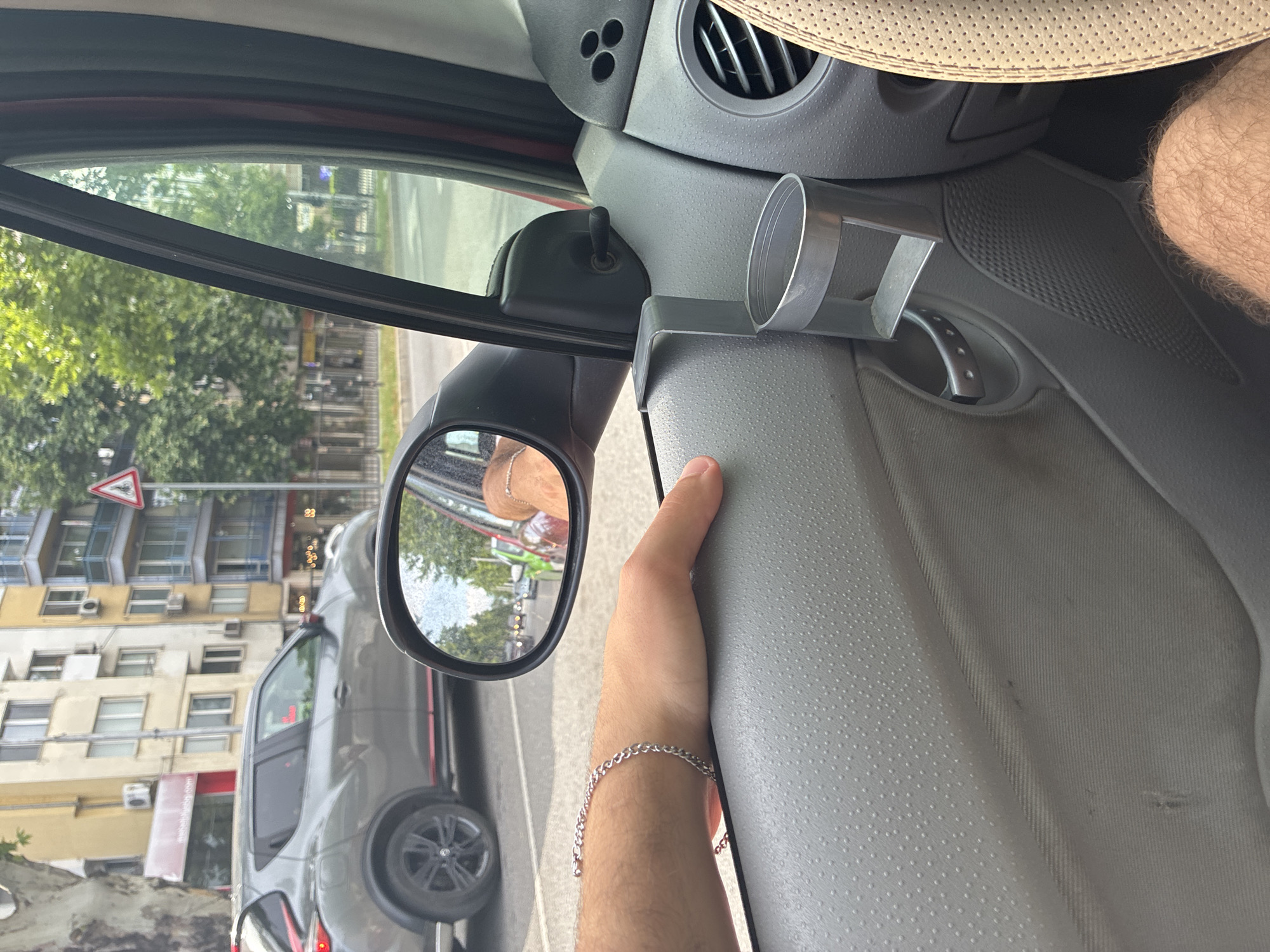 The image captures a first-person perspective from inside a car, looking outwards from the passenger side towards an urban street in Plovdiv, Bulgaria. The hairy right arm of a person, wearing a silver chain bracelet, rests casually on the textured grey car door panel. A section of the person's hairy leg is also visible further up, suggesting the person is seated in the car.
Inside the vehicle, a grey, cylindrical cup holder is attached to the door panel, positioned below an air vent and a small black control switch. A section of a beige, perforated car seat cover is visible in the upper right. The side mirror reflects parts of the street, another vehicle, and a reflection of a person's head, possibly wearing a hat.
Outside the car, a street scene unfolds under bright, sunny daytime conditions. The background features several multi-story buildings, likely residential or commercial, characterized by balconies and numerous air conditioning units. One building displays a red sign with the text "dabakh.bg," while another has the number "33" visible. Green trees line the street. A grey car, resembling an SUV, is prominently visible alongside or behind the observer's vehicle. A red and white triangular yield sign is positioned near the buildings. The image captures a moment of travel or pause within a city environment.