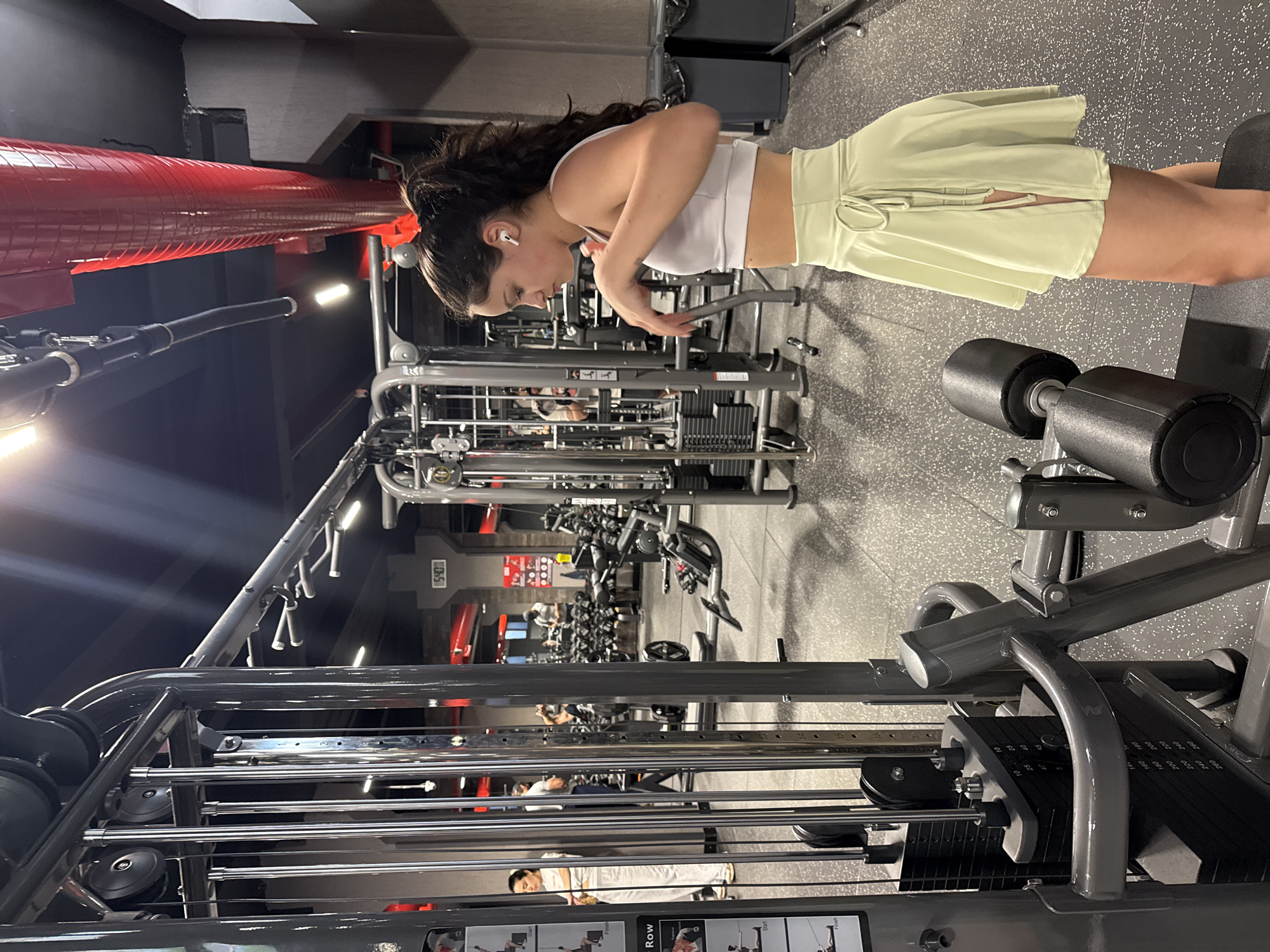 The photograph, taken indoors, depicts a woman in a fitness center. The woman, wearing a white sports bra and a short, light green skirt, has her hair tied back. She is facing a weight machine, seemingly stretching or preparing to use it. In the background, there are rows of other gym equipment and blurred figures of people, suggesting the facility is moderately populated. The gym features black ceilings, red pipes, and a dark gray floor. The presence of machinery indicates a modern, well-equipped gym environment. The location in the city of Yerevan, Armenia cannot be determined from the image.