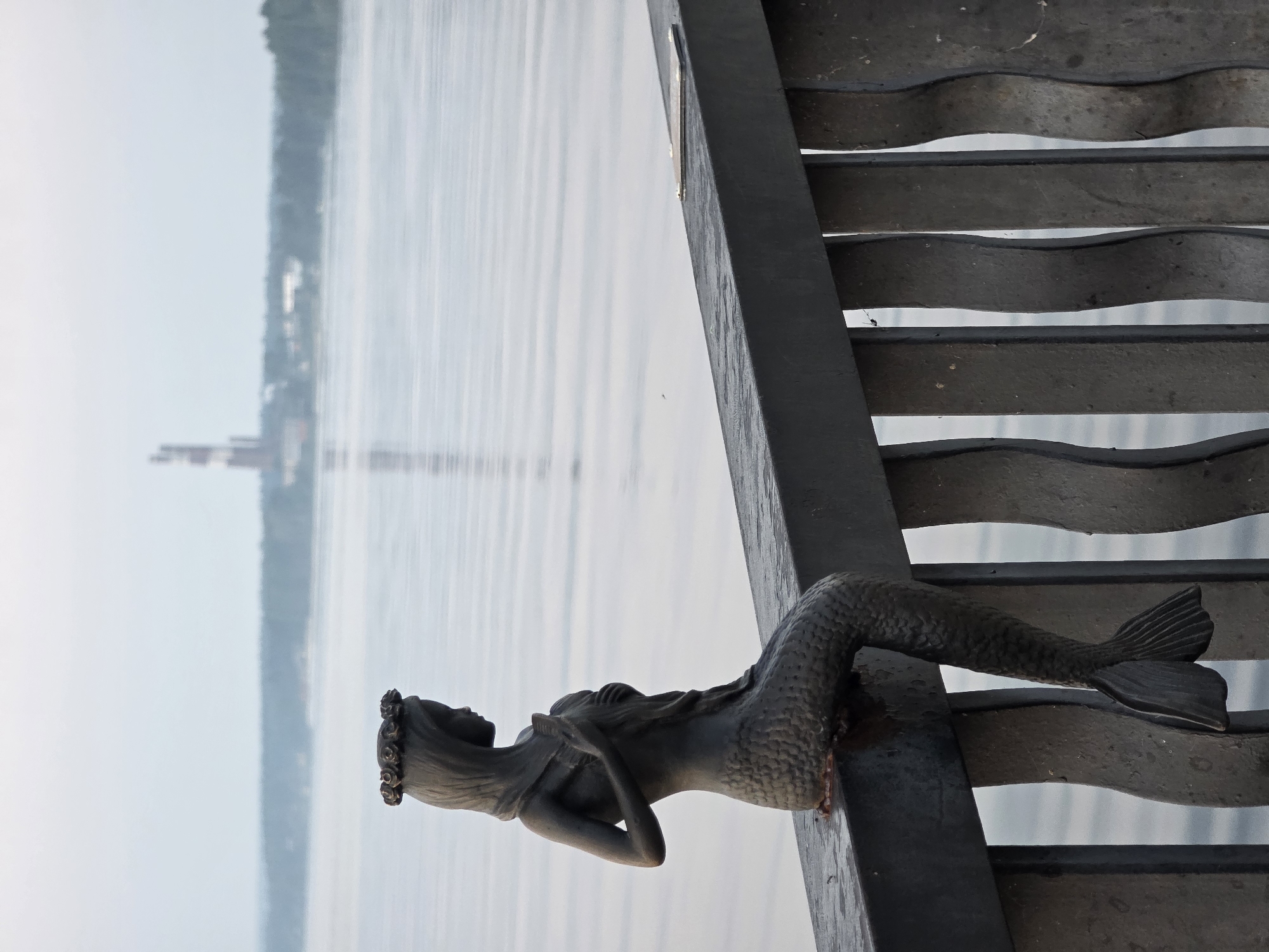 A bronze sculpture of a mermaid is perched on a dark metal railing overlooking a wide river. The mermaid is depicted with her upper body visible, adorned with a floral wreath on her head and holding her hands to her chest. Her lower body transitions into a scaled tail that hangs over the railing. The river is calm with gentle ripples, and a hazy shoreline with trees and a distant industrial structure, likely a power plant with chimneys, is visible across the water. The railing is made of black metal with decorative, wavy vertical supports. The overall atmosphere is serene and slightly overcast. This sculpture is located on the embankment of the Kama River in Chaikovsky, Russia.