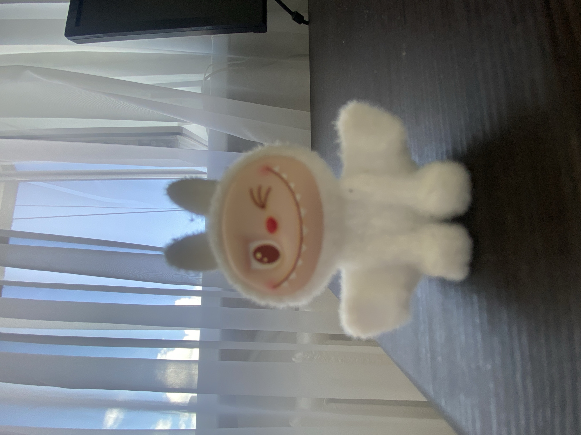 A white, fluffy plush toy with large, round ears and a smiling face with one eye winking and a red nose is positioned in the foreground of the image. The toy is sitting on a dark, textured surface, likely a desk. Behind the toy, sheer white curtains are drawn across a window, revealing a bright blue sky with some clouds visible. To the right of the toy, partially visible, is a black computer monitor. The lighting suggests it is daytime. The image was taken in Uchaly, Russia.