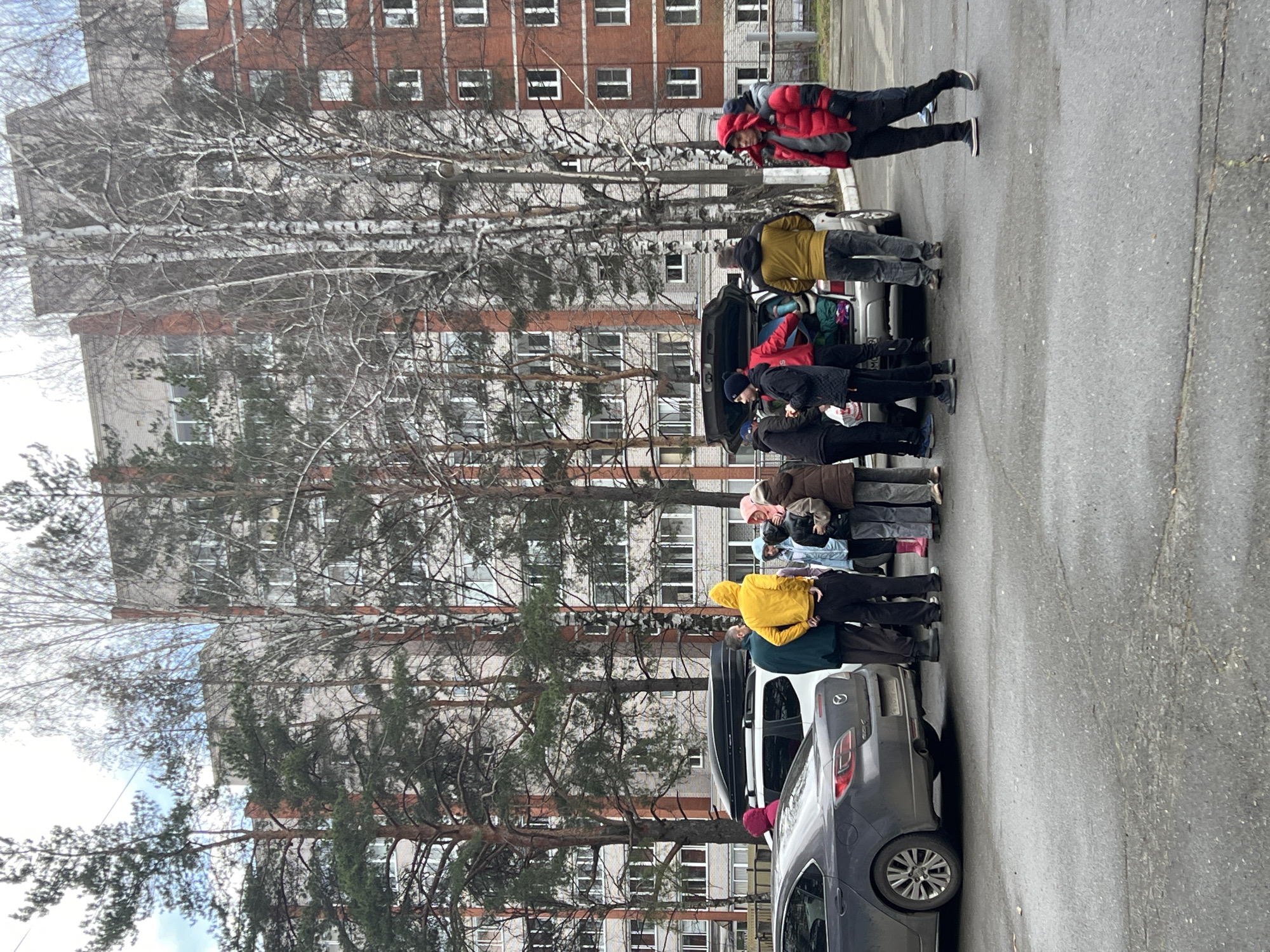 The image captures an outdoor scene in Chaikovsky, Russia, depicting a group of people gathered around parked cars on an overcast day. Approximately eight to ten individuals, dressed in warm winter or early spring clothing such as heavy jackets, coats, and hats, are visible.
The central activity appears to involve loading or unloading items from the trunk of a silver hatchback or SUV. Luggage, bags, and blankets are visible within the open trunk, suggesting preparation for travel, a move, or a group outing. Another dark grey sedan or hatchback is parked closely, featuring a roof box, which further implies preparations for a journey or transportation of gear.
In the background, a multi-story apartment building, characterized by its red brick sections and lighter-colored facades, stands prominently. Its architecture suggests a typical residential structure. Between the paved street or parking area and the building, several bare deciduous trees, possibly birches, and evergreen pine trees are interspersed, indicating late winter or early spring conditions. The ground is a cracked asphalt surface.
The weather appears cool or cold, as suggested by the heavy attire of the people and the bare branches of the deciduous trees. No visible text can be clearly identified on any objects or signage within the frame. The scene conveys a sense of practical activity or transition rather than leisure.