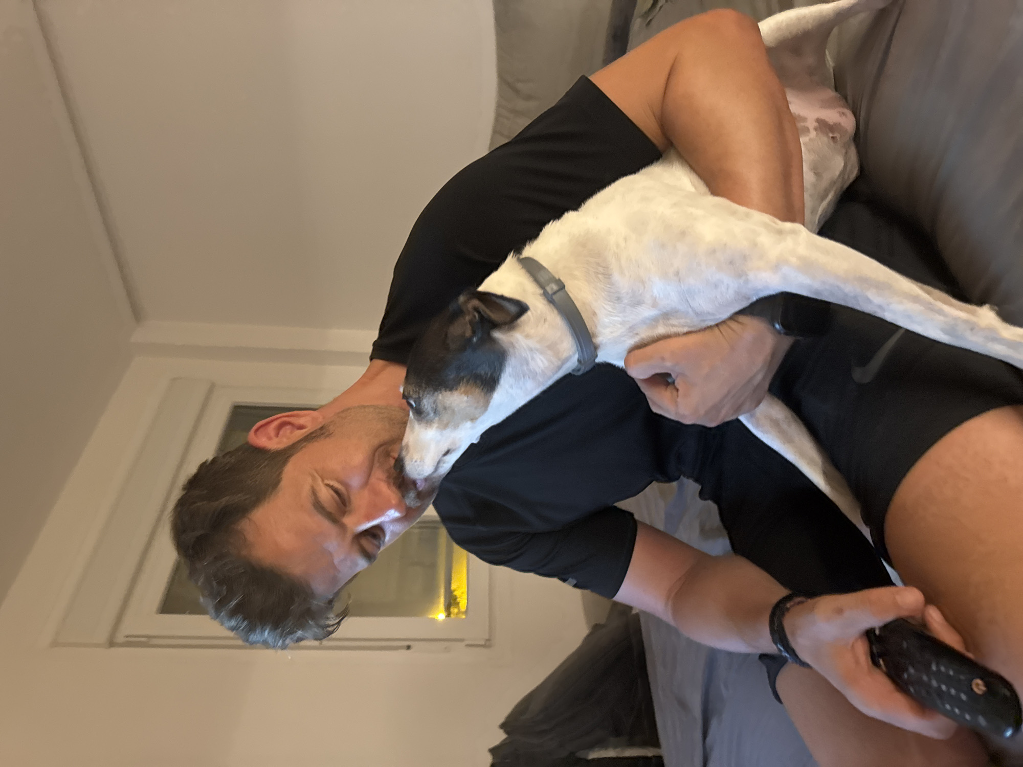 A man with short, brown hair and a mustache is holding a white and black dog. The dog is licking the man's face. The man is wearing a black t-shirt and black shorts. He is sitting on a couch with a remote control in his left hand. The background is a room with a window and a bed. The image appears to be taken indoors, likely in a residential setting in Murcia, Spain, given the implied context.