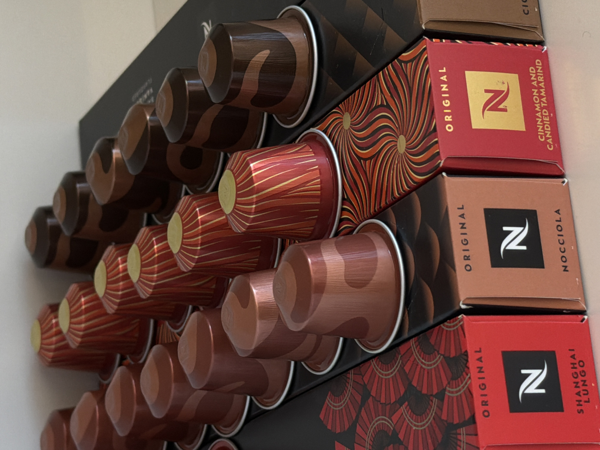 The image shows a collection of Nespresso coffee capsules and their corresponding packaging. Four different varieties are visible: Cioccolatino, Cinnamon and Candied Tamarind, Nocciola, and Shanghai Lungo. The capsules are displayed in rows, partially inserted into the packaging boxes. The location in Riga, Latvia is not evident from the image.