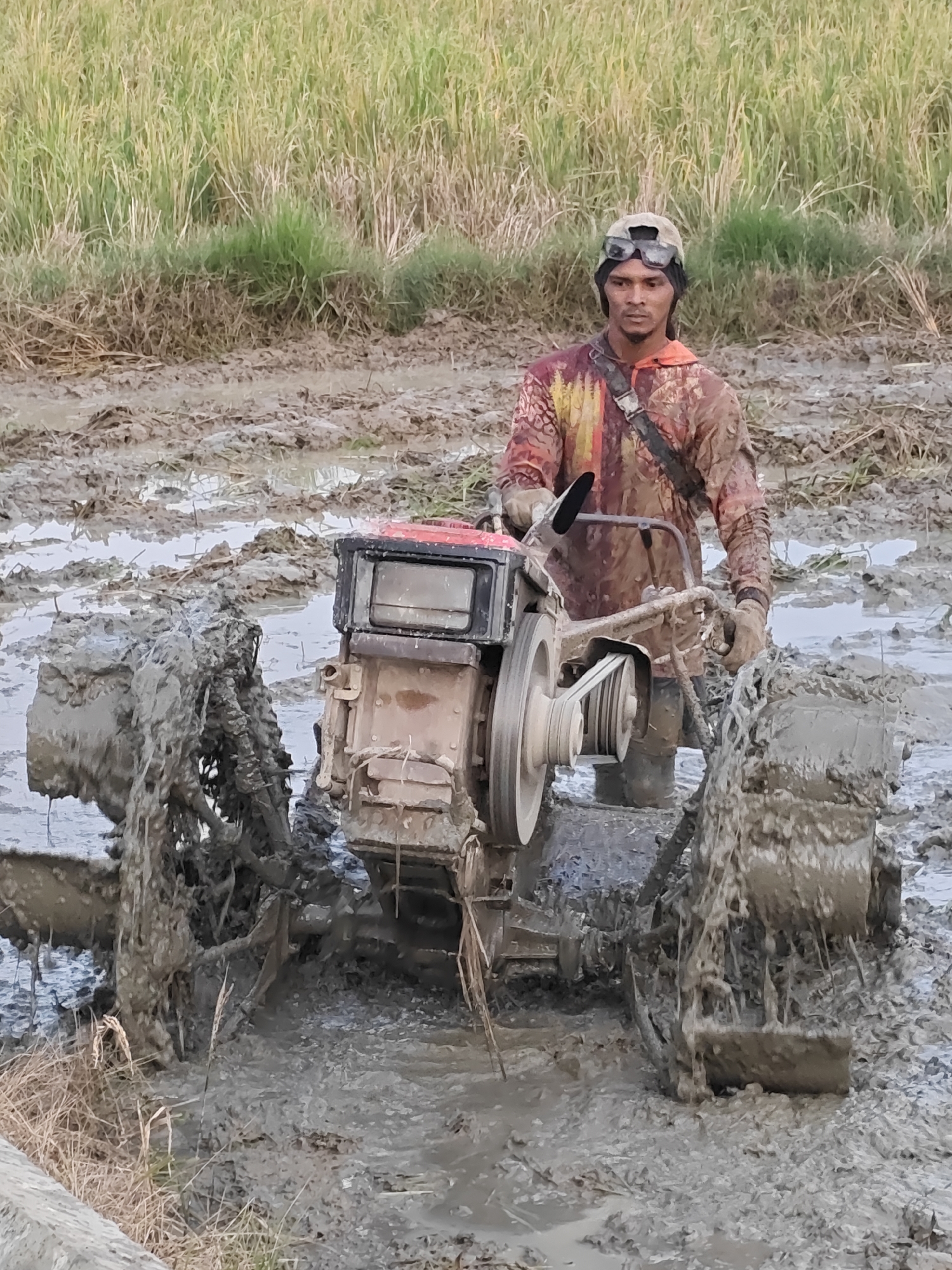 In Meunasahalue, Indonesia, a man operates a tiller in a muddy rice paddy. The man, wearing a patterned long-sleeved shirt, sunglasses, and a cap, is partially submerged in the water and mud. The tiller, a red and silver machine, is caked in mud, with its wheels deep in the paddy. The environment is characterized by a vast, muddy field with tall green rice stalks in the background. The lighting suggests it is daytime.