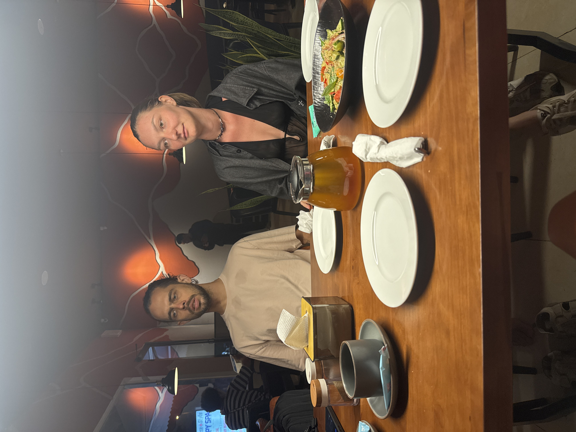 A couple sits at a wooden table in what appears to be a restaurant. The woman, wearing a black top and a grey denim jacket, sits across from the man who wears a light beige long-sleeved shirt. They are sitting next to each other as they are eating their meal. The table is set with white plates, a black serving dish containing a colorful salad, a tea set, and a tissue box. The wall behind them is decorated with a mural that shows a silhouette of the city.
The reflection on the wall is from the restaurant's interior, displaying other customers. The room lighting appears to be a warm hue. Based on the location and the style of the mural, it is plausible that this scene is taking place in a restaurant in Đà Nẵng, Vietnam, although it cannot be confirmed with certainty from the image alone.
