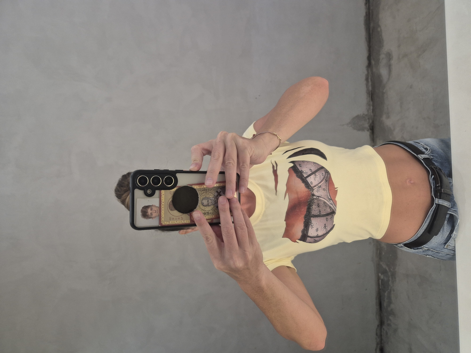An individual is captured taking a mirror selfie indoors, holding a smartphone with both hands. The person is wearing a light yellow short-sleeved crop top featuring a graphic print of a bra on the front, revealing their midriff. They are also dressed in blue jeans secured with a black belt. A gold-colored bracelet is visible on their right wrist.
The smartphone has a black case with three prominent camera lenses on the upper left side. A circular black pop socket is attached to the back of the phone. On the phone's case, beneath the pop socket, there's a visible decorative element that appears to be a golden rectangular image, possibly resembling currency or an ornate design, which includes what looks like a face or a stylized figure and some indistinct script. A reflection of the person's head, showing some hair, can be faintly seen in the phone screen.
The background behind the person is a textured light gray wall, possibly concrete or a similar material, with a darker vertical line or corner visible on the right side of the frame. There is no clearly legible text on the clothing or the phone case, beyond the general appearance of script on the golden element. The activity depicted is a casual mirror selfie, typical of indoor settings. The provided context indicates the location is Dubai, United Arab Emirates, although no specific visual cues from the image itself confirm this.