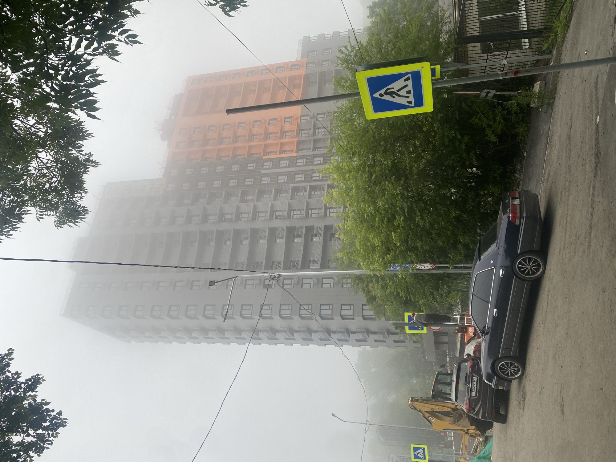 In a foggy urban environment, a multi-story apartment building under construction dominates the background. The building's facade is a mix of gray and orange sections, with visible balconies. In the foreground, a dark sedan is parked on a wet asphalt road. To the left, a yellow excavator is partially visible, suggesting ongoing construction. Pedestrian crossing signs with blue backgrounds and white pedestrian symbols are placed along the roadside. A lamppost stands near one of the signs. Green trees and foliage are scattered throughout the scene, partially obscuring the lower parts of the building and the street. A person is visible on a ladder near a lamppost, possibly working on it. The atmosphere is hazy, indicating poor visibility.