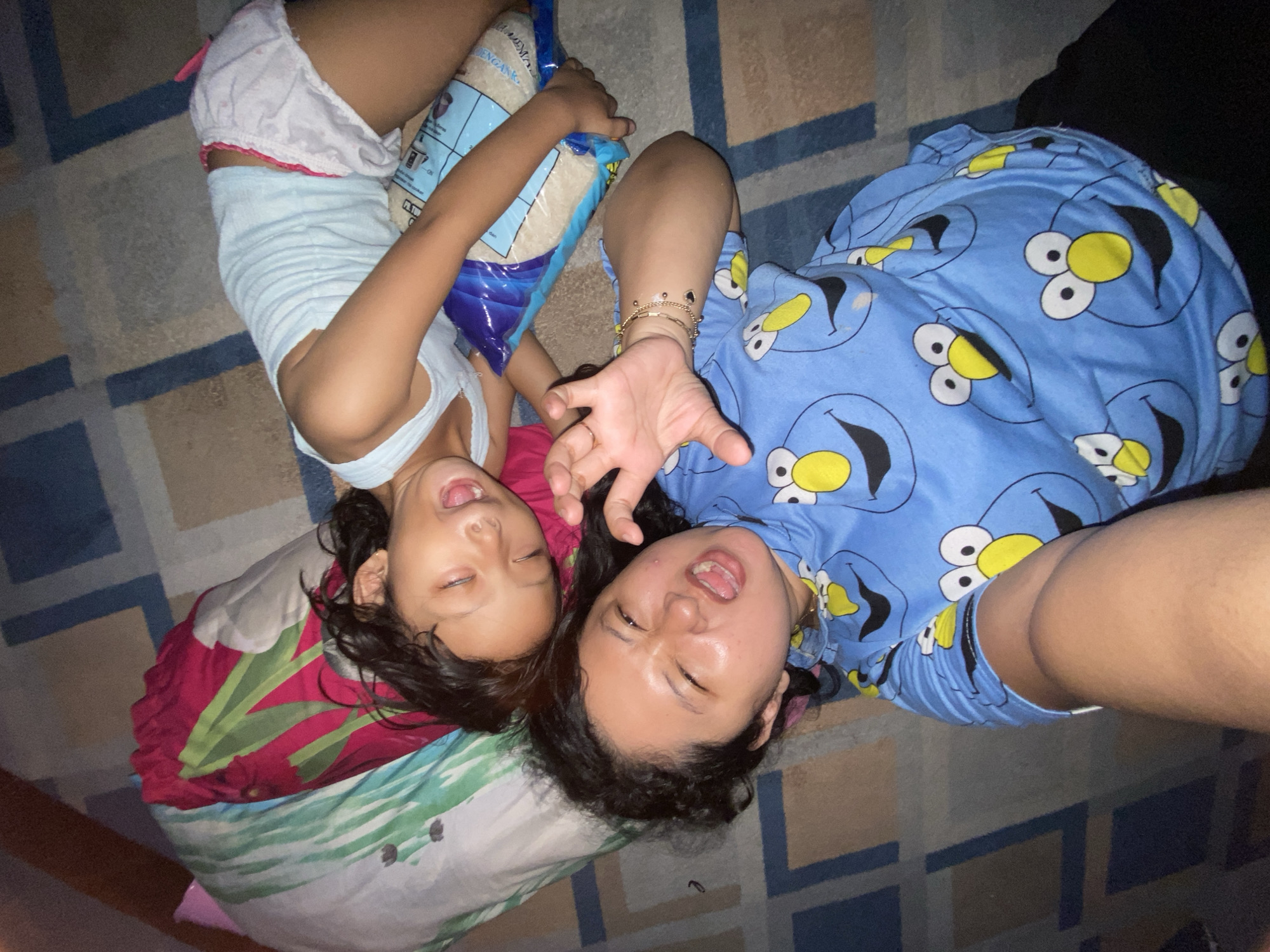 The image depicts two individuals lying on a patterned rug. One individual, presumably a child, is wearing a light blue tank top and white shorts with red trim, and is holding a blue bag. The other individual, likely an adult, is wearing a blue shirt with Cookie Monster patterns. Both individuals have their mouths open and appear to be looking at the camera. The location is assumed to be in Gelam, Indonesia, based on the user's instruction. Other details include the patterned rug and a partially visible object near the child's head.