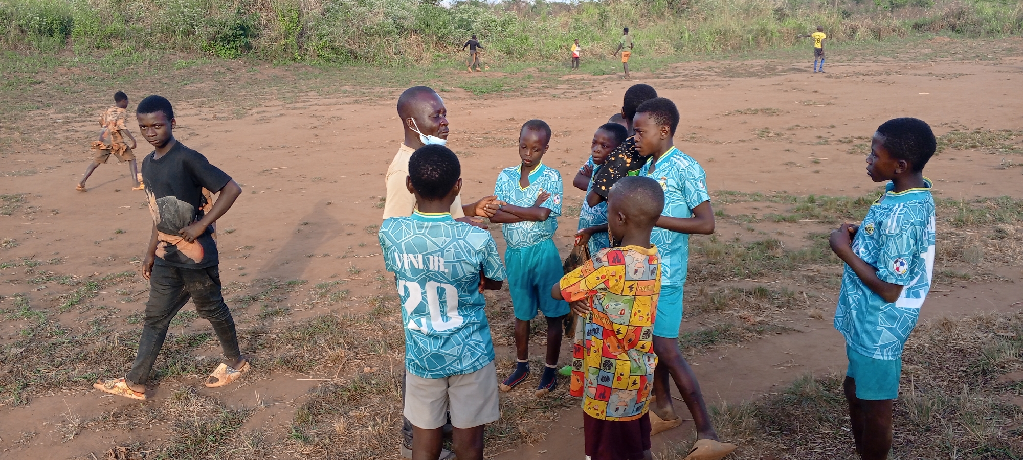 This outdoor photograph captures a gathering of children and an adult male on a wide, dusty dirt field in Ngalula, Democratic Republic of the Congo. The ground is uneven, characterized by dry earth with sparse patches of grass, and is bordered by thick green vegetation in the background under what appears to be a clear, late afternoon sky.
In the midground, an adult male with dark skin, wearing a light brown t-shirt and a light blue face mask pulled below his chin, stands conversing with a group of approximately seven children. Most of the children are dressed in matching light blue or turquoise football jerseys featuring a geometric pattern, along with matching shorts. One child, seen from behind, has "JUNIOR." written above the number "20" on the back of their jersey. Another child in the group wears a distinctive yellow t-shirt with a colorful, multi-patterned design, depicting what look like cartoon characters.
To the left, a boy in a black t-shirt, dark trousers, and patterned slip-on sandals looks directly towards the camera while walking. In the background, other children are visible across the expanse of the field, some appearing to be engaged in play. One child in the far background is seen wearing a yellow jersey with the number "19" on the back. The scene suggests a community sporting event or practice session, with the adult seemingly coaching or instructing the young players. The children appear attentive, indicating an ongoing conversation or lesson.