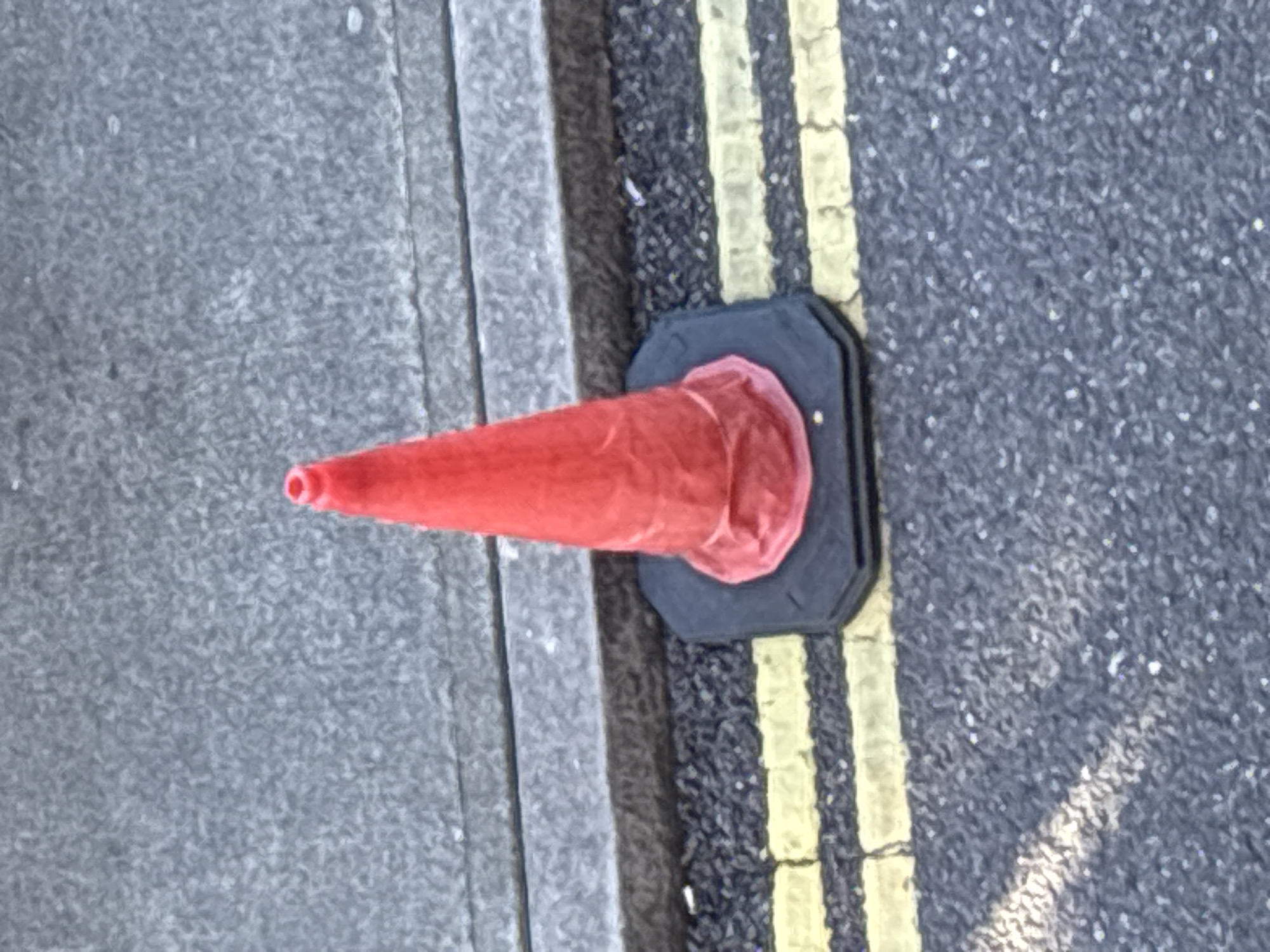 A single, bright red traffic cone stands on a grey asphalt road, positioned just beside a raised concrete curb. The cone's base is black and octagonal, and it is slightly wrinkled, suggesting it might be made of a flexible material. Two parallel yellow lines are painted on the asphalt in front of the cone, indicating a road marking. The background consists of more grey asphalt and the textured surface of the concrete curb. The scene appears to be an outdoor urban environment, likely a street. Without further context or discernible landmarks, it is impossible to pinpoint a specific location within the city of Cork, Ireland. There are no people, actions, or interactions visible in the image.