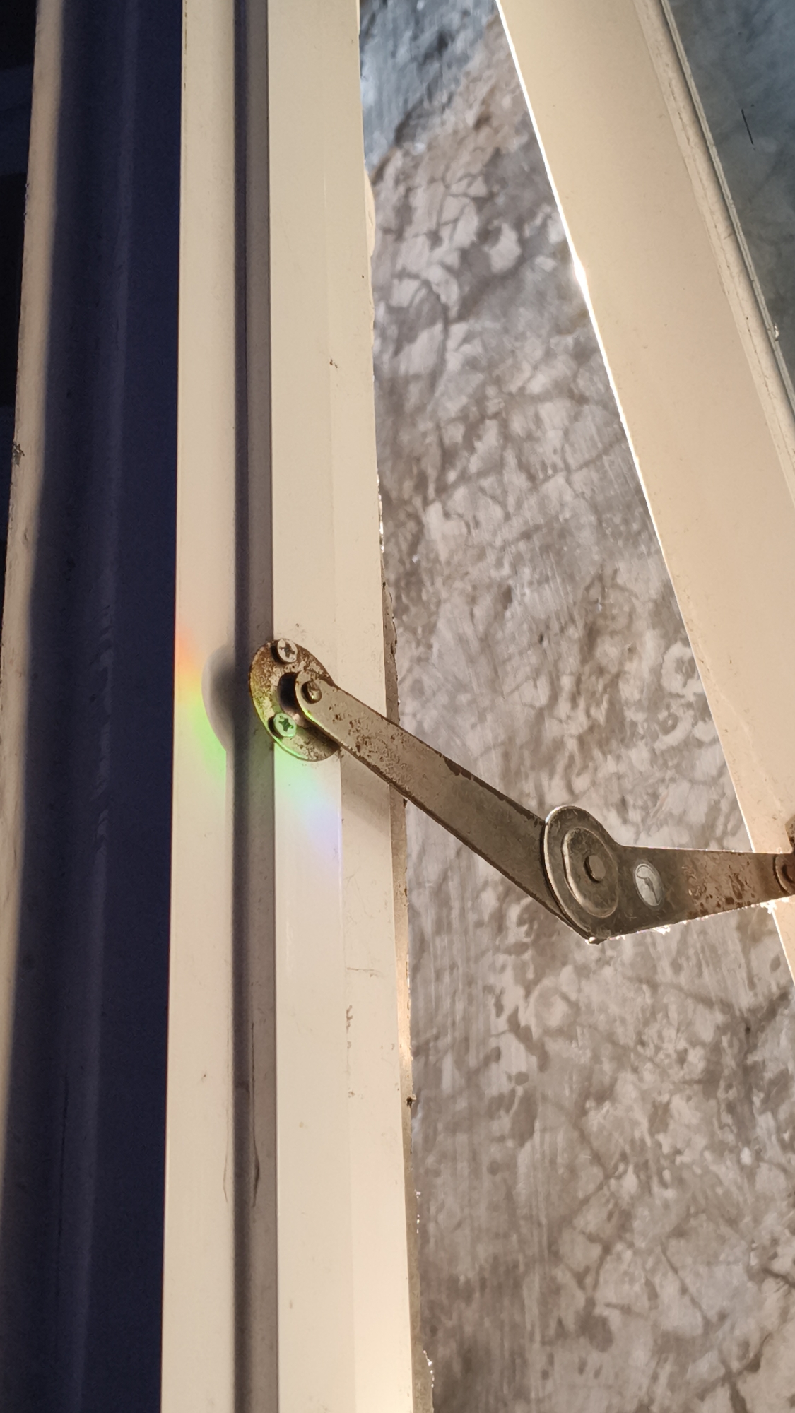 The image shows a close-up of a white window frame with a rusted metal hinge. A rainbow-like refraction of light is visible on the left side of the window frame. The background appears to be a textured concrete wall. The scene is indoors, likely a residential or commercial building. There are no people or animals visible in the image. The image does not provide any information about its location in Kerep Kidul, Indonesia.