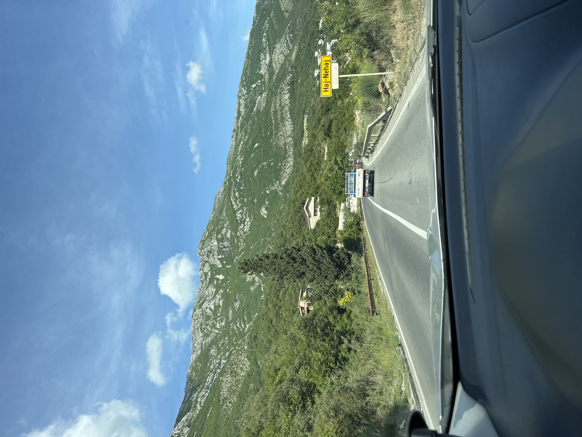 The image depicts a view from inside a vehicle traveling on a road near Đurmani, Montenegro. A yellow sign for "Haj-Nehaj" is visible on the right side of the road. A mountain covered in vegetation rises on the left. Two vehicles are ahead on the road. The sky is blue with some clouds.