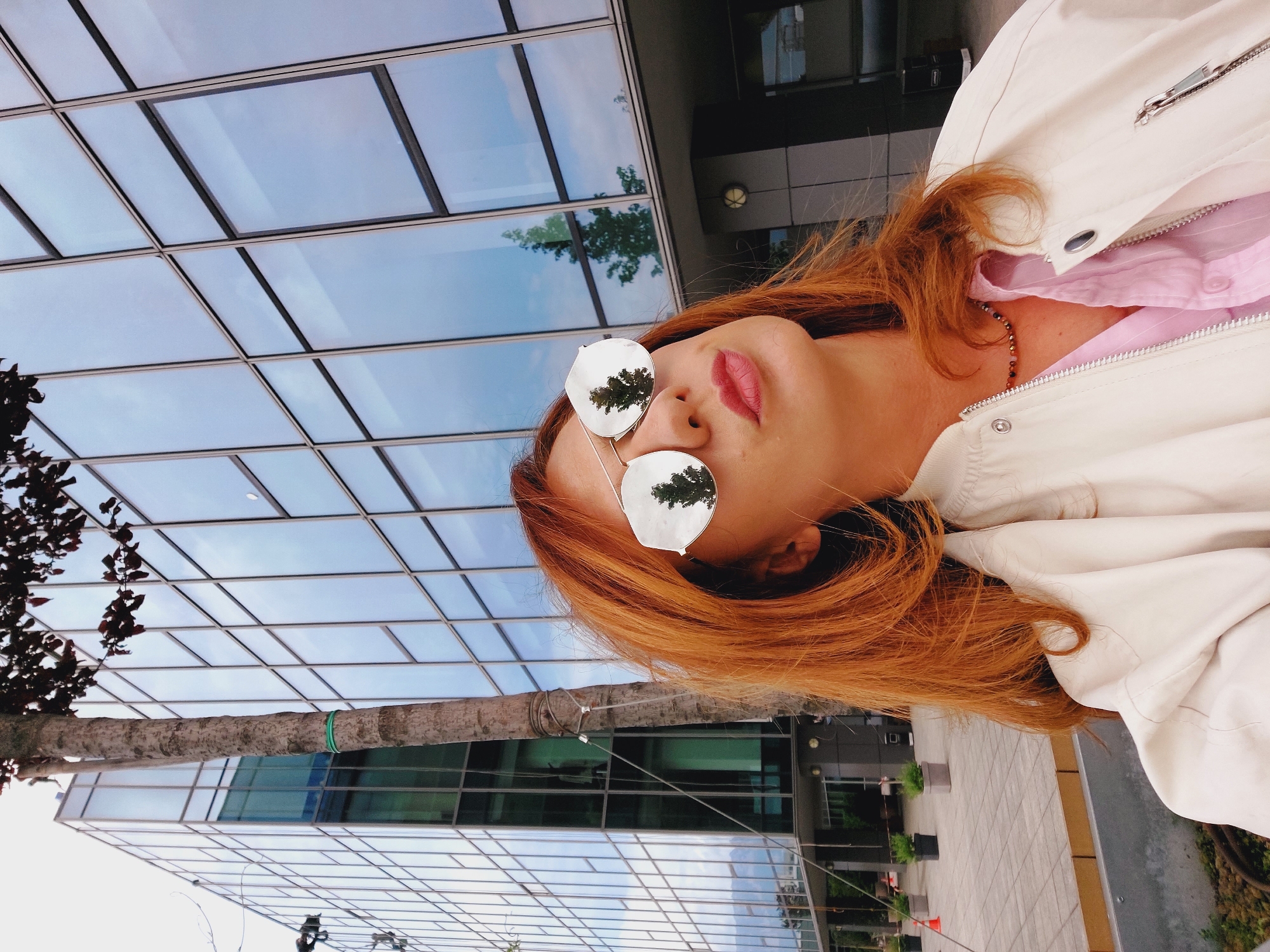 A woman with auburn hair is taking a selfie outdoors in front of a modern glass-fronted building. She is wearing a light-colored bomber jacket over a pink collared shirt and a beaded necklace. Her sunglasses have mirrored lenses that reflect trees and the sky. The background shows the facade of a large office building with many windows, a tree trunk in the foreground, and paved ground with planters. The scene appears to be in an urban environment, likely Sofia, Bulgaria, given the architectural style and vegetation.