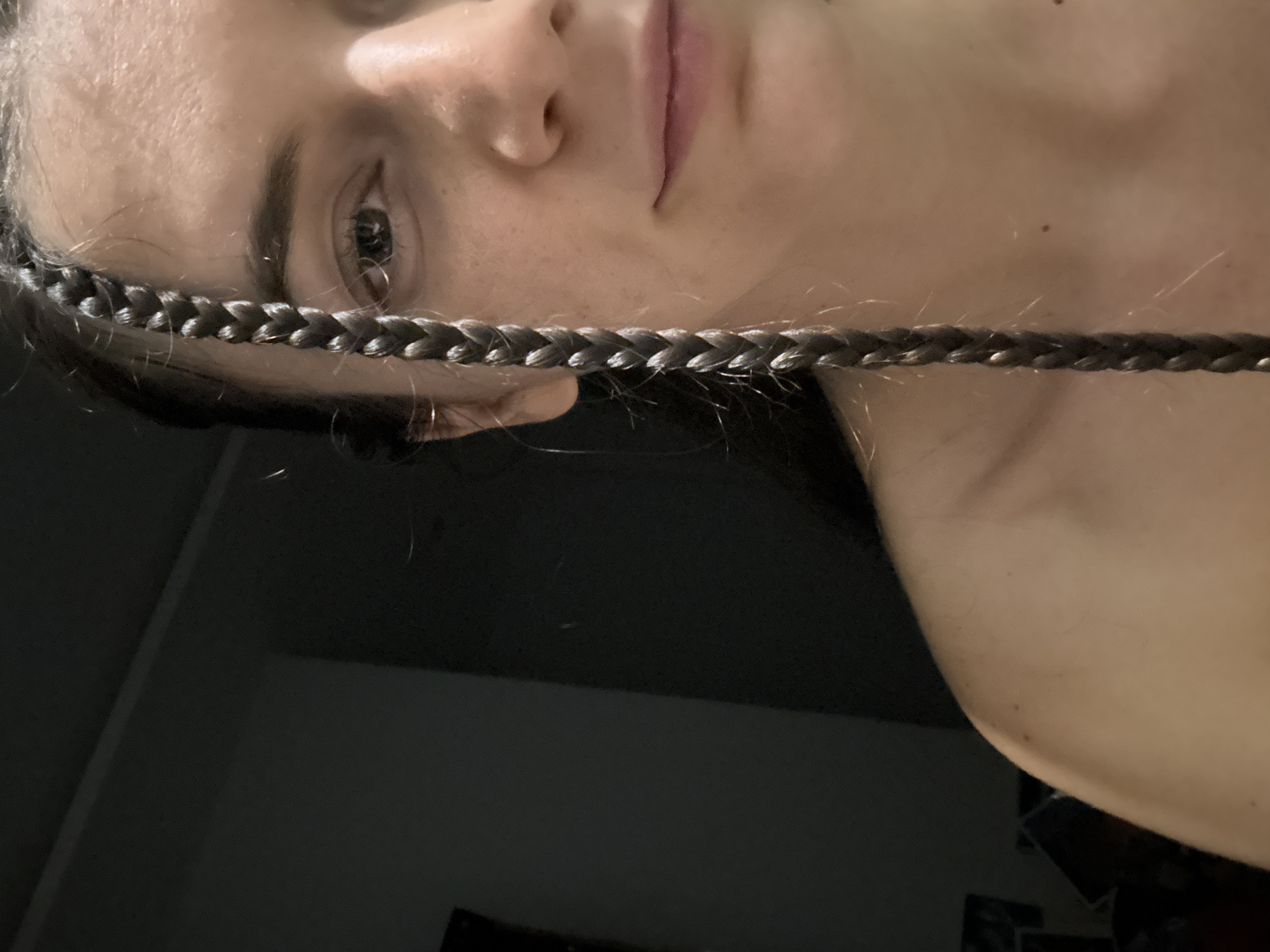 This close-up portrait features a person, likely female, positioned with her head tilted 90 degrees to the left. A prominent, dark braided section of hair stretches horizontally across the lower half of her face, extending from just below her left ear across to the right side of the frame. Her left eye is open and looks directly forward, displaying a neutral or contemplative expression. The bridge of her nose and part of her upper lip are visible above the braid.
The lighting is soft and appears dim, casting parts of the background into shadow. Her skin tone is light, and fine, lighter flyaway hairs are visible around her face and along the braid. The background is largely dark and undifferentiated, suggesting an indoor setting with minimal light, possibly showing a wall or dark furniture. No specific objects, patterns, or text are discernible in the environment. The visible area of her shoulder or chest appears bare, blending into the dark background. The image evokes a sense of quietude and focus on the subject's gaze. This media file was captured in Mũi Né, Vietnam.