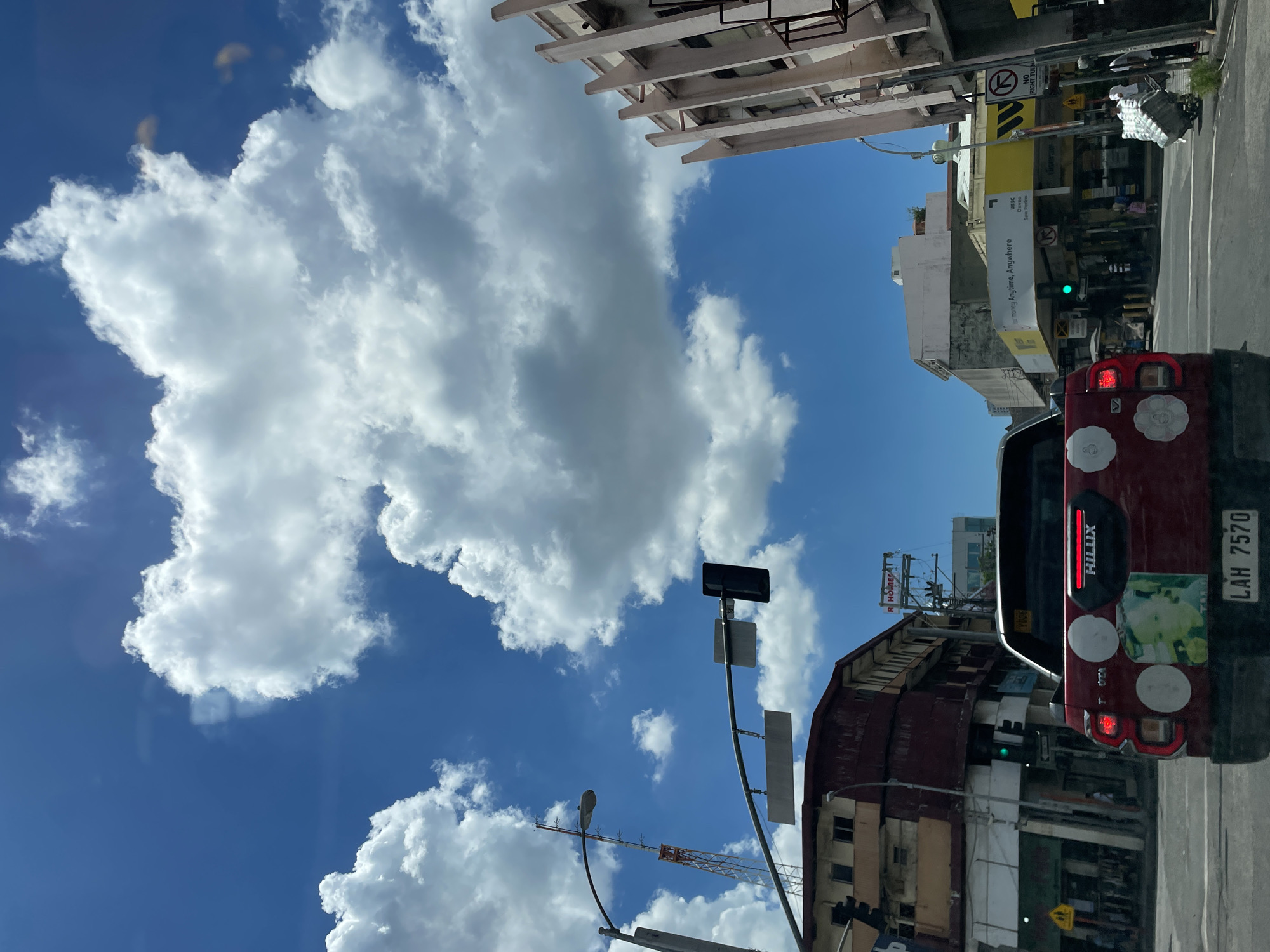 The image captures a street view in Davao City, Philippines, looking forward from a vehicle. A bright blue sky with fluffy white clouds dominates the upper portion of the frame. In the foreground, the rear of a red Toyota Hilux pickup truck is visible, adorned with several circular white stickers and a larger square sticker on the tailgate. The truck's license plate reads "LAH 7570". The vehicle is stopped at an intersection, indicated by a functioning traffic light showing a green signal.
The surrounding environment reveals a bustling urban setting. On the left, older buildings with weathered facades line the street, one appearing to be red and another with a faded yellow exterior. A tall communication tower with orange and white sections stands prominently in the distance. On the right side of the street, modern buildings with a distinctive architectural style, featuring vertical concrete elements, are visible. A yellow awning with signage for "USC Davao San Pedro" stretches across a commercial establishment. Pedestrians are present on the sidewalks, and a person is seen pushing a cart laden with goods. Another building with a white and light-colored facade is situated further down the street. The scene suggests a typical day in a city center, with traffic and pedestrian activity.