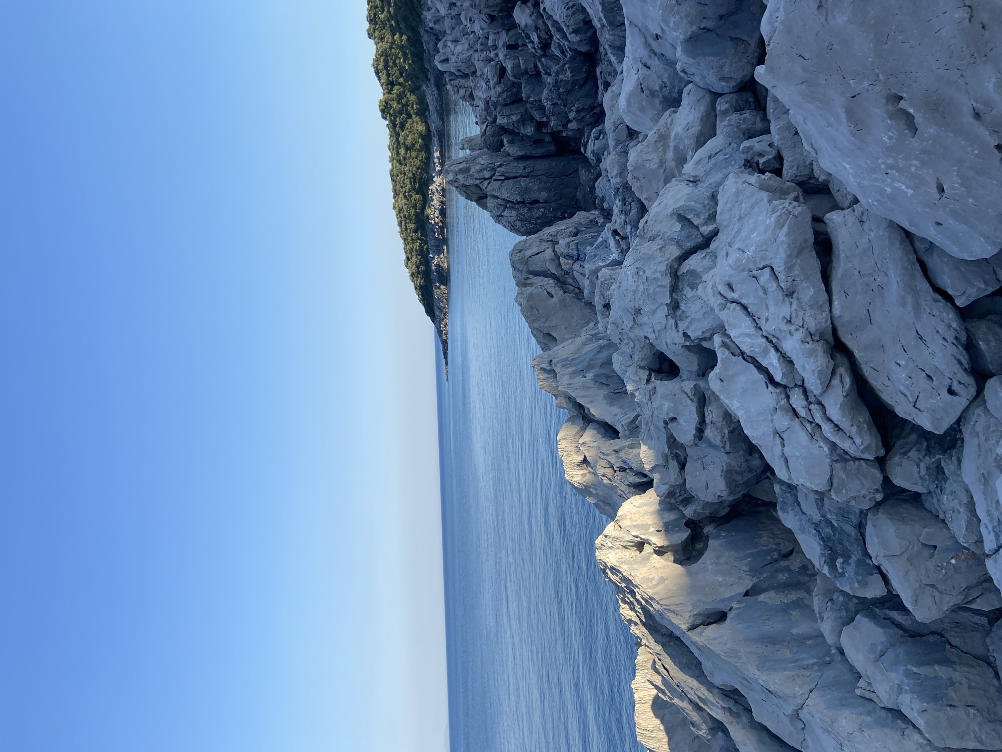 The image depicts a rocky coastline meeting a calm sea under a clear blue sky. The foreground is dominated by a jumble of large, grey rocks, some casting shadows that suggest the sun is at an angle, likely indicating morning or late afternoon. To the left, the expansive sea stretches towards the horizon, its surface smooth with subtle ripples. In the distance, a green, tree-covered slope rises from the coastline, hinting at land with vegetation. The overall scene evokes a sense of tranquility and natural beauty. There are no people, objects, or visible text in the image.