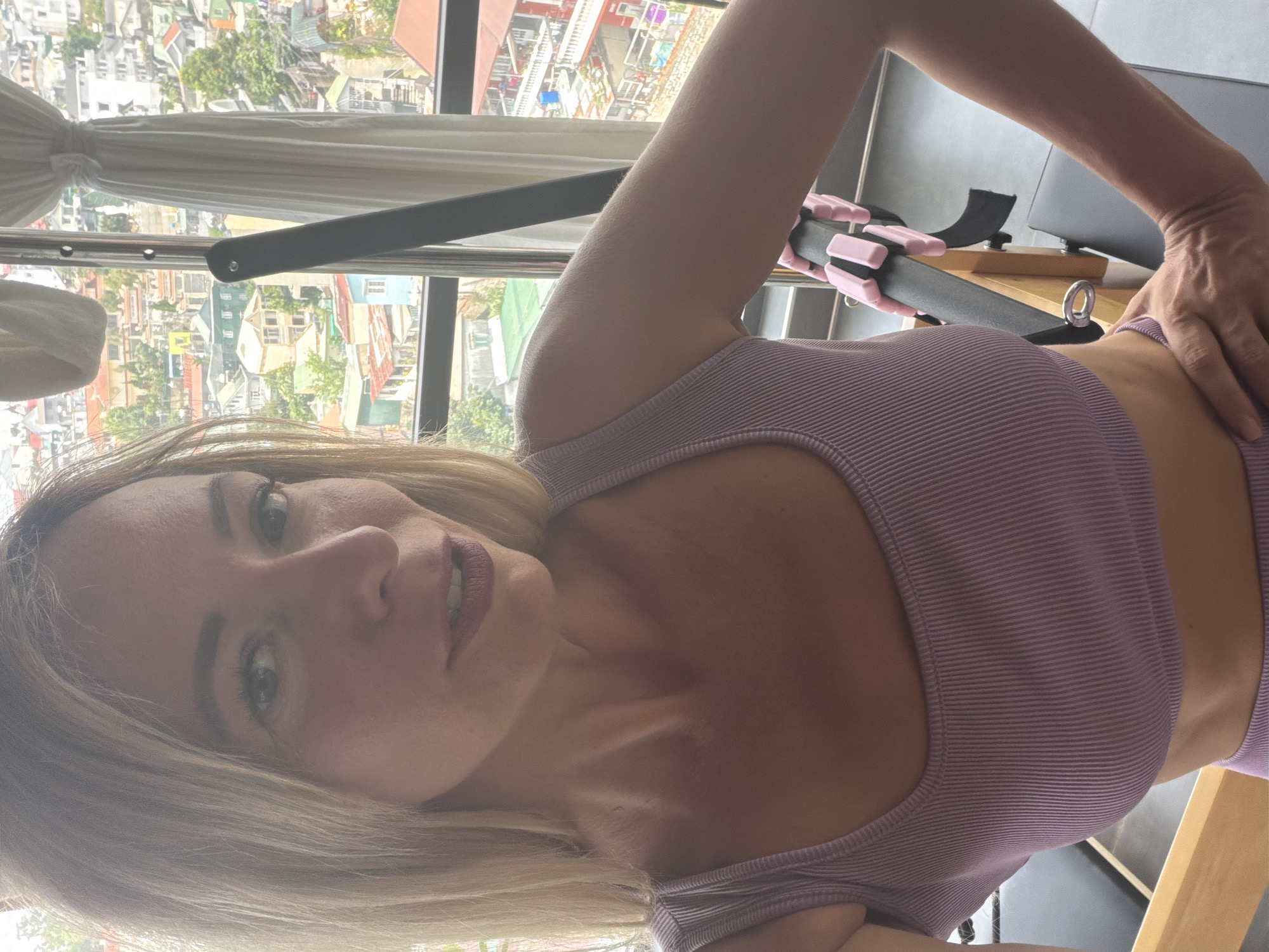 A woman with blonde hair and light-colored eyes, wearing a purple ribbed crop top, takes a selfie in front of a window overlooking a dense urban area. The buildings are a mix of colors, with red-tiled roofs and some with balconies. Trees are visible interspersed among the structures. The woman is positioned to the left of the frame, with her right arm extended towards a piece of fitness equipment with pink accents. A white curtain hangs to the right of the window. The scene suggests an indoor setting, possibly an apartment or gym, with a view of Da Lat, Vietnam.