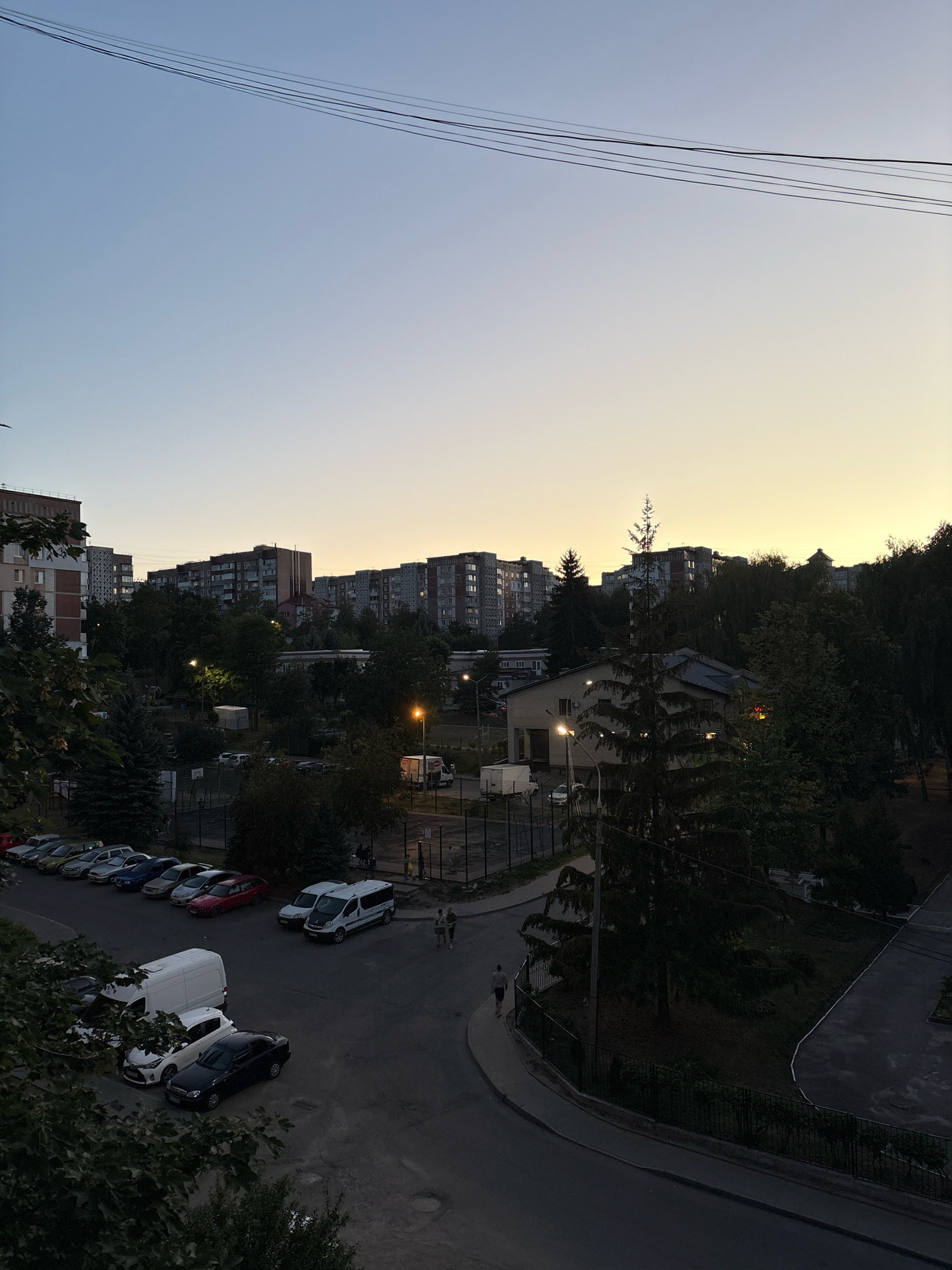 An elevated, full-frame, outdoor, eye-level, daytime shot captures a residential area in Ternopil, Ukraine, during dusk. The sky transitions from a pale blue at the top to a soft yellow near the horizon. Overhead, multiple dark electrical wires are strung across the frame. Below, a series of apartment buildings, characterized by their uniform, blocky architecture, dominate the midground. Between the buildings, dense foliage and trees create a green buffer. In the foreground, a paved road curves into the scene, with parked vehicles along its edge. Several cars, a white van, and a white minivan are visible. A few individuals are walking on a sidewalk near the parked vehicles. The overall scene is quiet and depicts a typical urban residential environment.