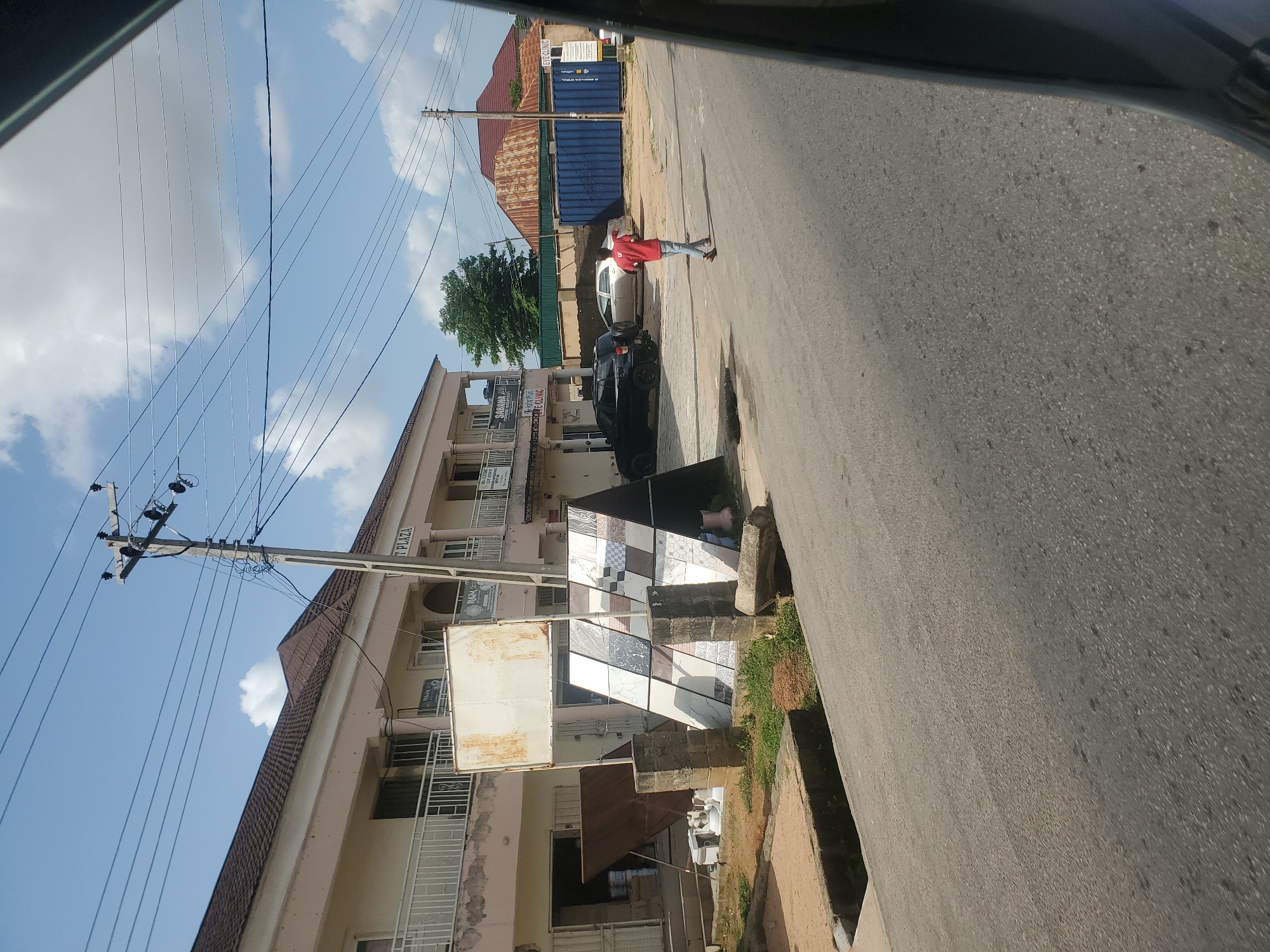 This image depicts a street scene in Kason, Nigeria. The foreground features an asphalt road with a concrete drainage channel on the left. In the mid-ground, a building with white and beige facade and balconies is visible. In front of the building, there is a display of ceramic tiles supported by brick pillars. A person in a red shirt and blue jeans walks away from the camera on the right side of the road. Behind the building, there are parked vehicles, including a black SUV and a silver car, and a large blue shipping container. The background shows a red-tiled roof, trees, and utility poles with overhead wires against a bright blue sky with scattered clouds.